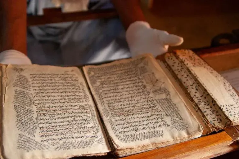 مخطوطة قديمة تُظهر نصوصاً عربية مكتوبة بخط يدوي، يُمسك بها شخص يرتدي قفازات بيضاء، تعكس التراث الثقافي لمكتبة عائلة الحبوط في شنقيط.