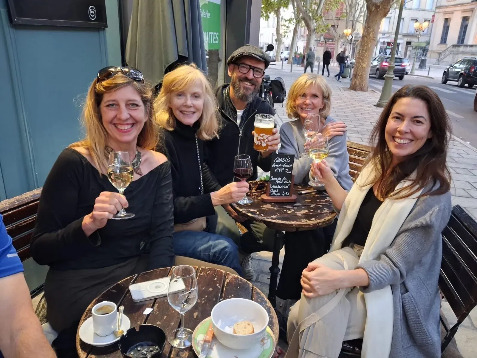 مجموعة من الأصدقاء يجلسون في مقهى في أوزيس، فرنسا، يحتفلون معًا بأكواب من النبيذ والبيرة، مع أجواء مريحة وودية.