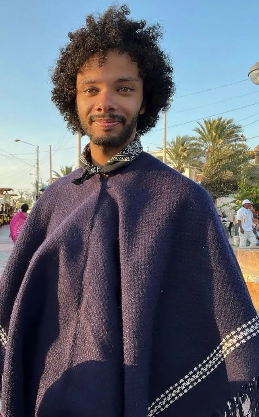 شاب يرتدي عباءة داكنة، يقف في منطقة مفتوحة تحت أشعة الشمس، مع أشجار النخيل في الخلفية. يعبر عن تجربته القاسية في السجون الفنزويلية.