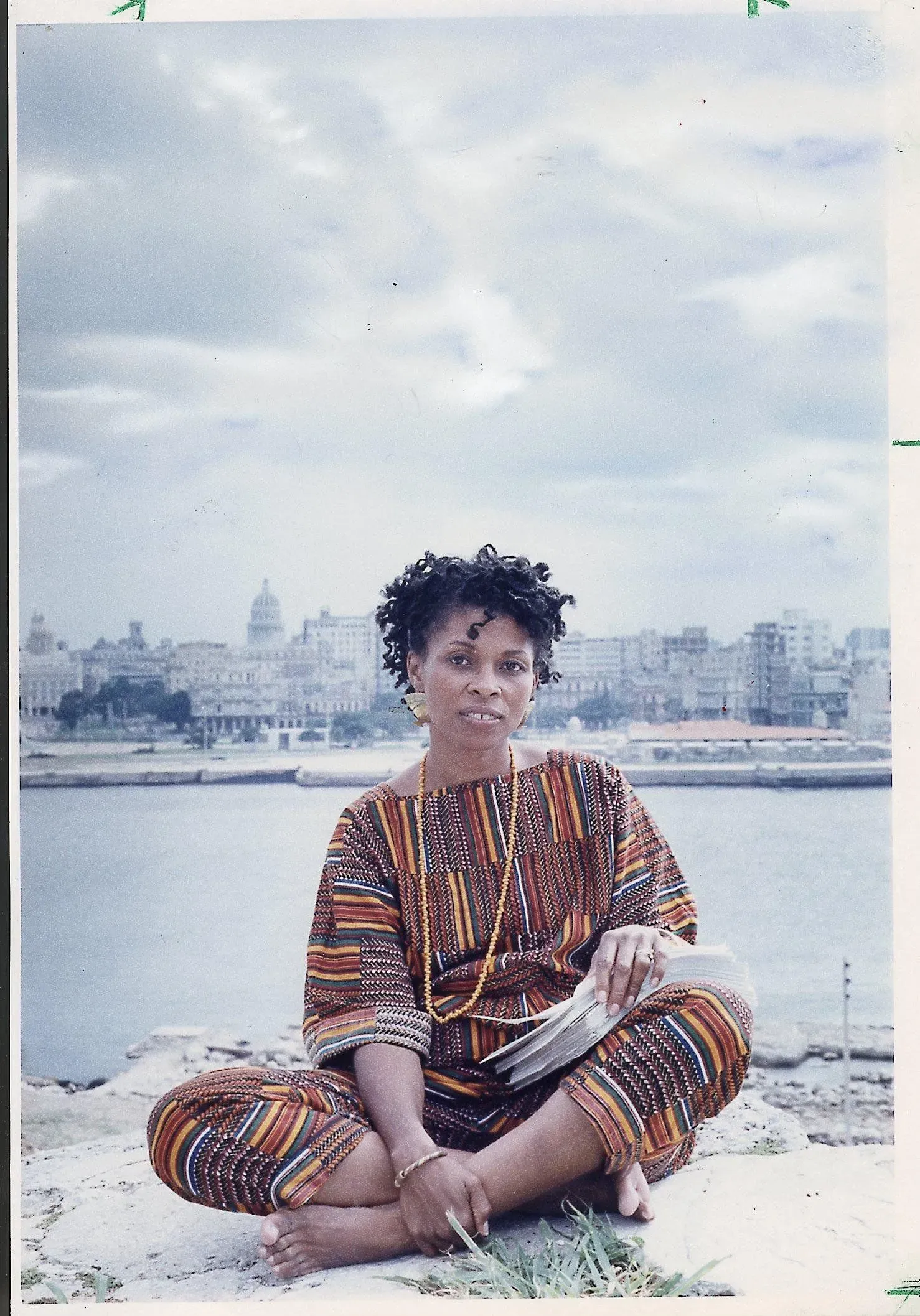 امرأة ترتدي ملابس ملونة تجلس على صخرة بجوار البحر في هافانا، مع معالم المدينة في الخلفية، تعكس روح المقاومة والثقافة.