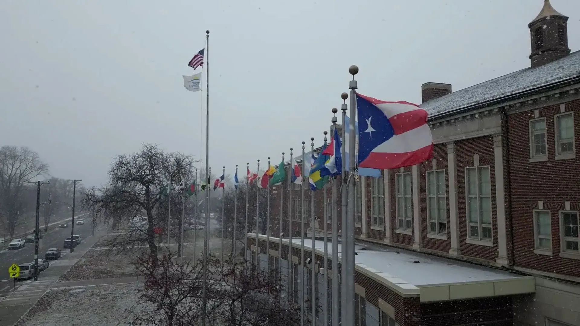 علم أمريكي وأعلام دولية تتمايل في الرياح تحت سماء ملبدة بالغيوم، مع تساقط الثلوج، أمام مبنى مدرسي في ديترويت.