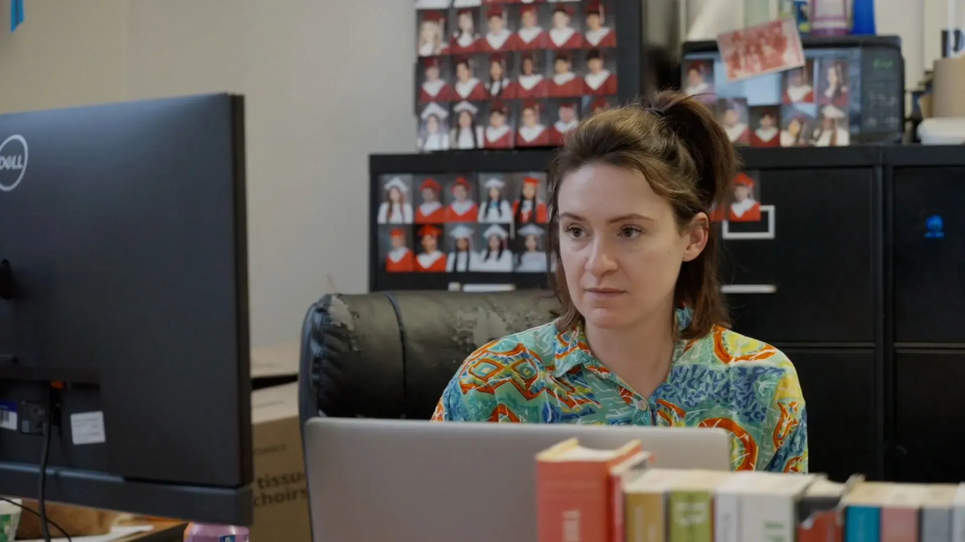 مدرسة تجلس أمام جهاز الكمبيوتر في فصل دراسي، محاطة بصور طلابها، تعكس تحديات التعليم ودعم الطلاب المهاجرين.