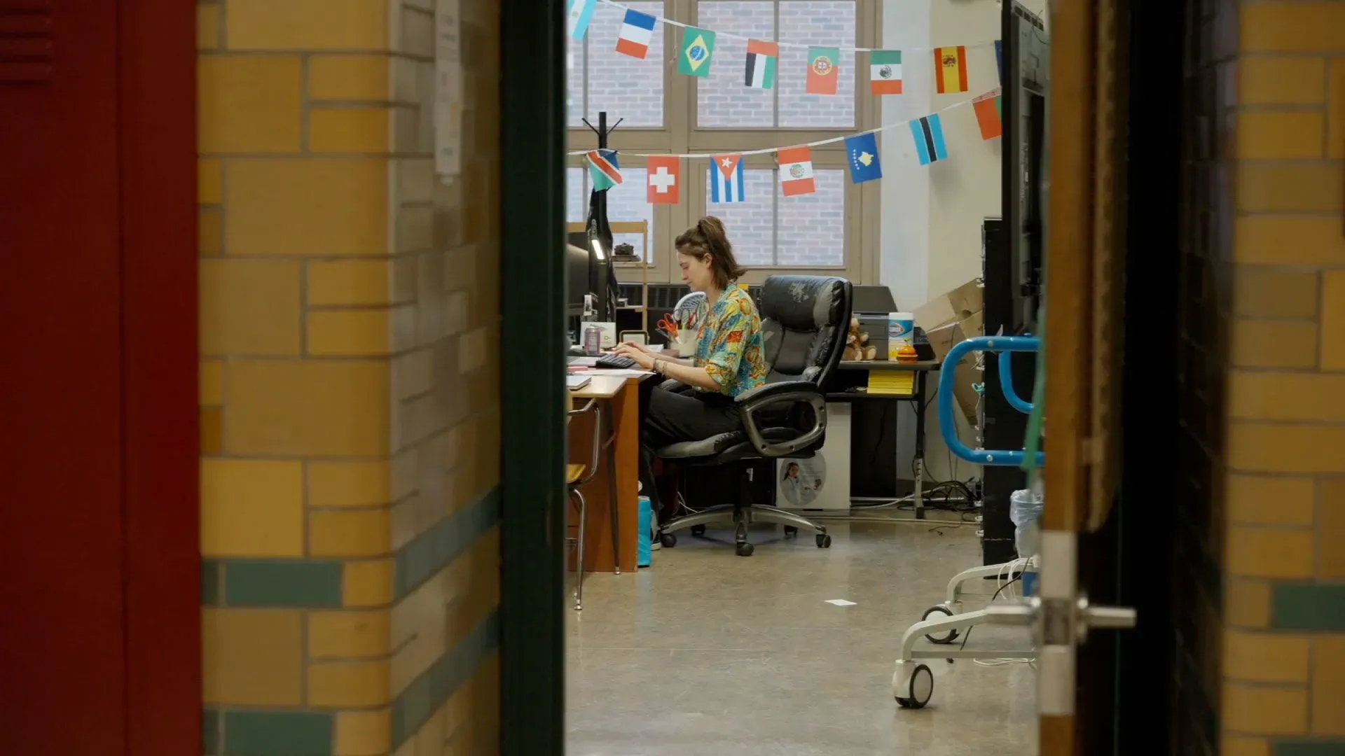 مدرسة تجلس في مكتبها داخل المدرسة، محاطة بأعلام دول متعددة، تعمل على دعم طلابها المحتجزين في مراكز الهجرة.