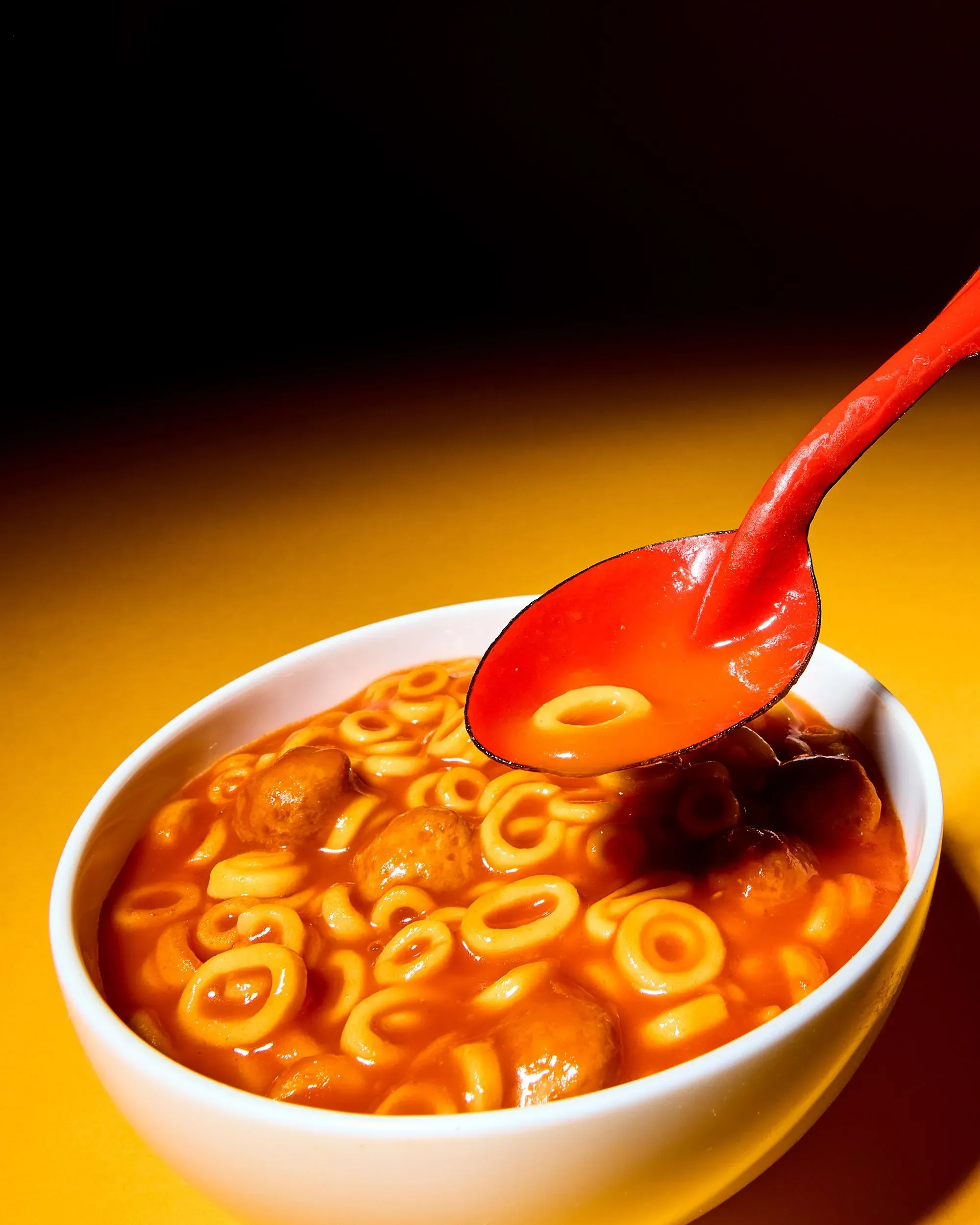 وعاء أبيض يحتوي على معكرونة حلزونية وكرات لحم في صلصة حمراء، مع ملعقة حمراء ترفع جزءًا من المحتوى، في خلفية صفراء.