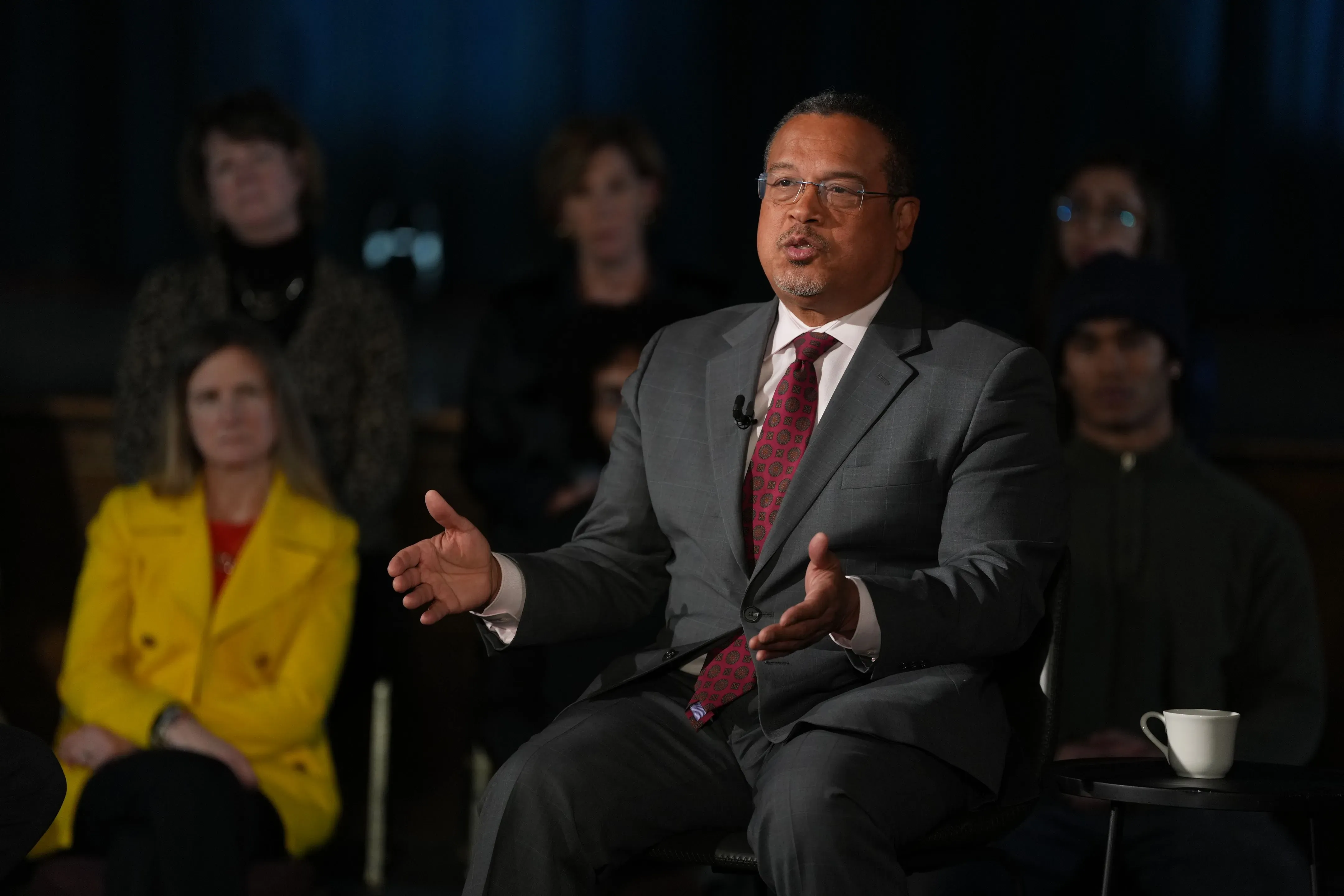 رجل يتحدث أثناء حدث عام في مينيسوتا، مع وجود جمهور خلفه. تعكس تعابيره قضايا الهجرة والتوترات المحلية.