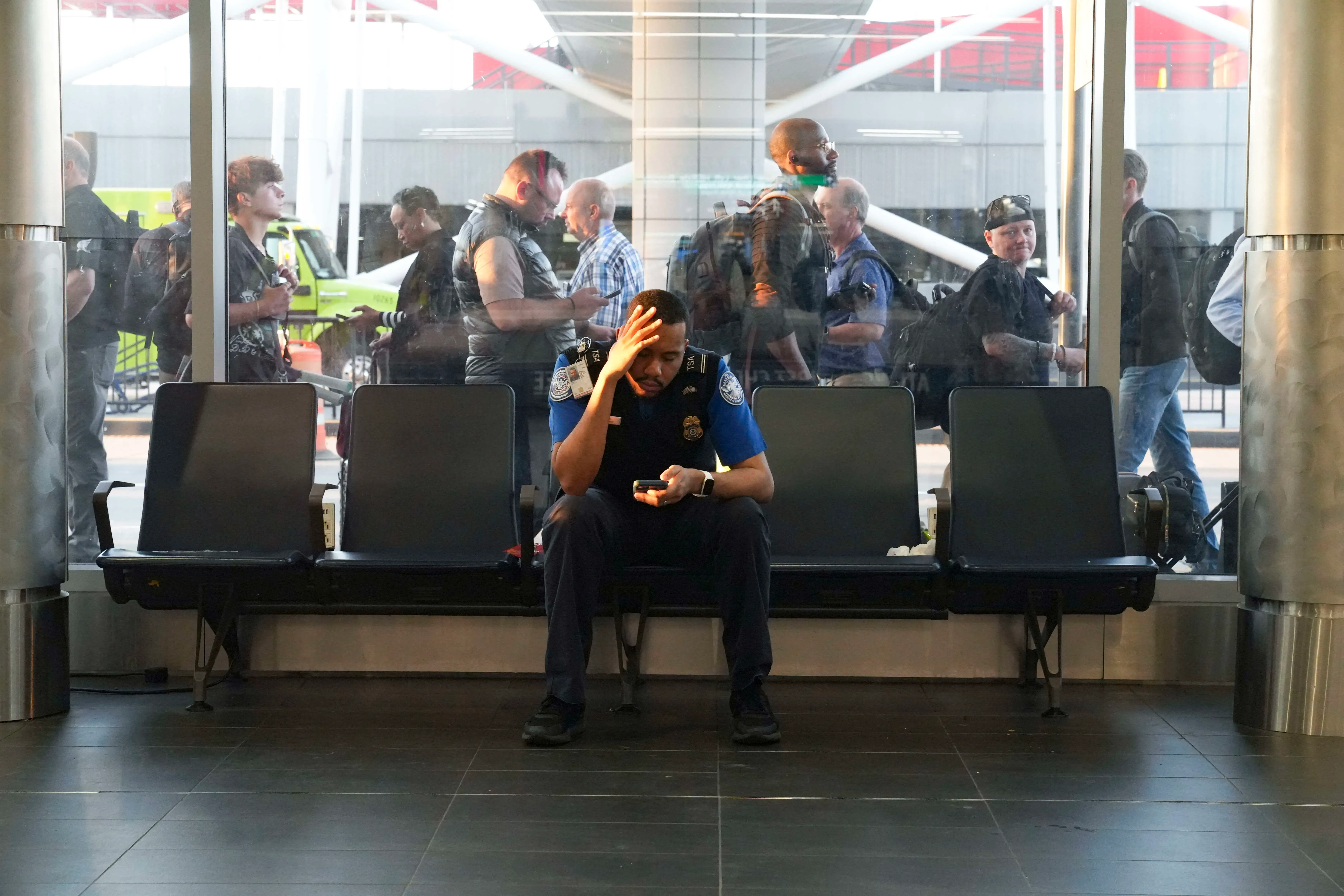 موظف من إدارة أمن النقل يجلس في مطار، يبدو عليه التعب والقلق، بينما ينتظر المسافرون في الطابور الطويل خلفه.