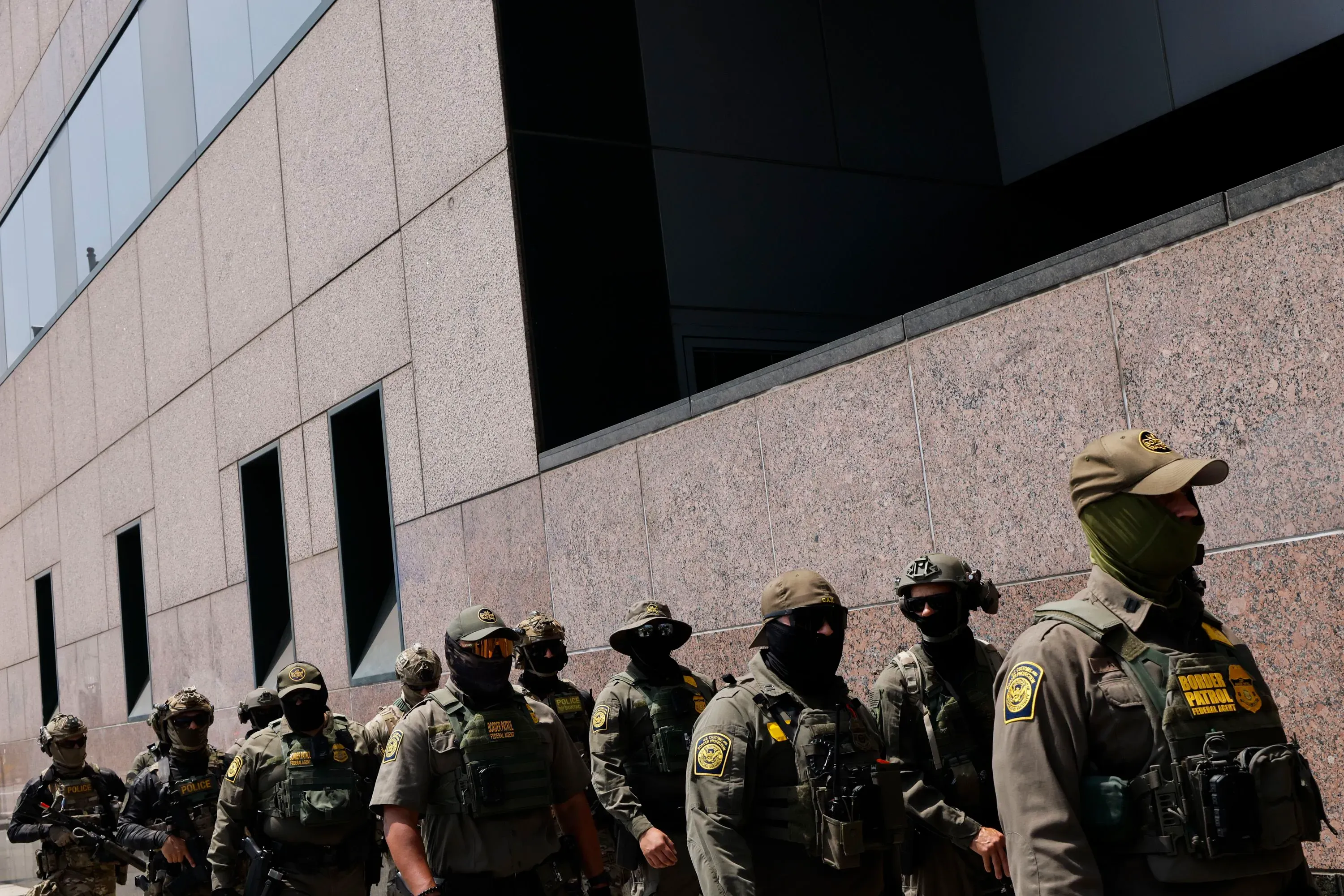 مجموعة من الضباط يرتدون زي إدارة الهجرة والجمارك، مع أقنعة ونظارات شمسية، أثناء تنفيذ عملية في منطقة حضرية.