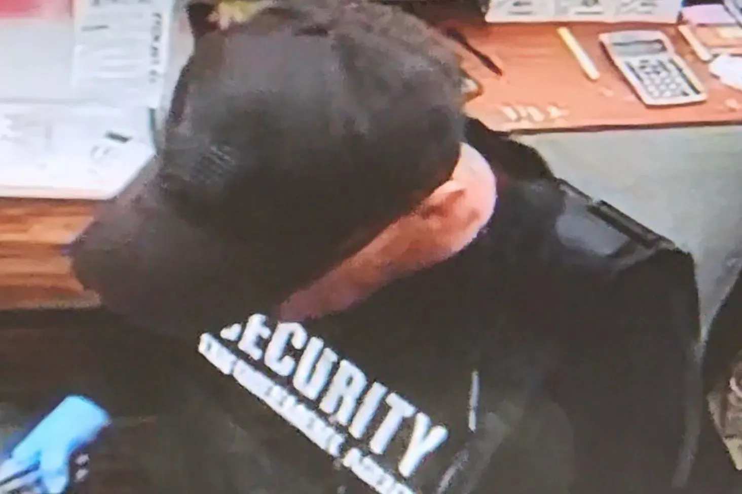 امرأة ترتدي سترة سوداء وقميصاً مكتوب عليه "SECURITY"، تخفي وجهها خلف قناع ونظارات شمسية بينما تتحدث مع موظف الفندق.