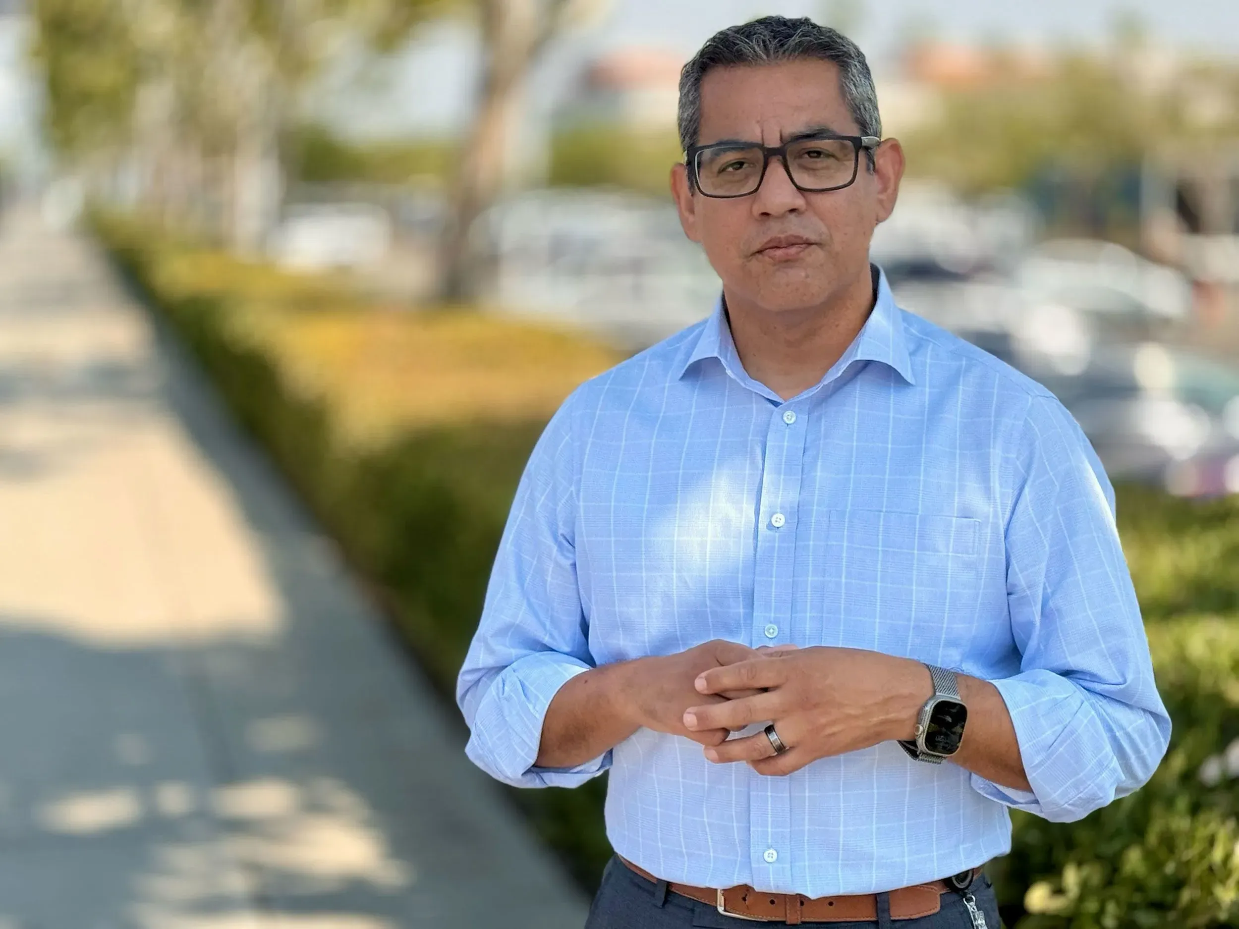 رجل يرتدي قميصًا أزرق ويقف في مكان عام، مع خلفية تحتوي على سيارات وأشجار، يتحدث عن قضايا انتحال شخصية ضباط الهجرة.