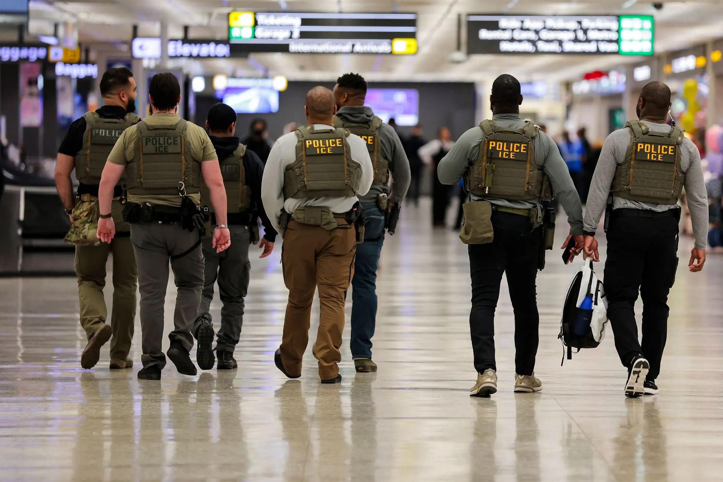 وجود مجموعة من عملاء إدارة الهجرة والجمارك يرتدون سترات تكتيكية في مطار، يوجهون المسافرين ويشرفون على الوضع الأمني وسط طوابير الانتظار الطويلة.