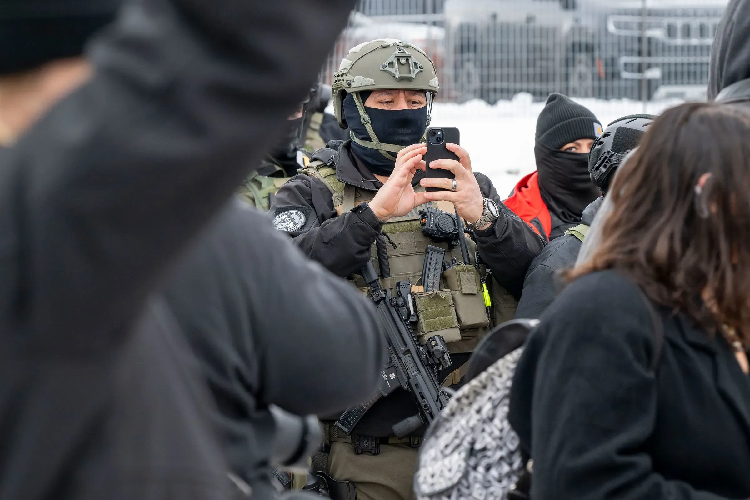 عميل من إدارة الهجرة والجمارك يرتدي زيًا عسكريًا ويستخدم هاتفه المحمول لتوثيق الأحداث أثناء عملية ميدانية، محاطًا بمحتجين.