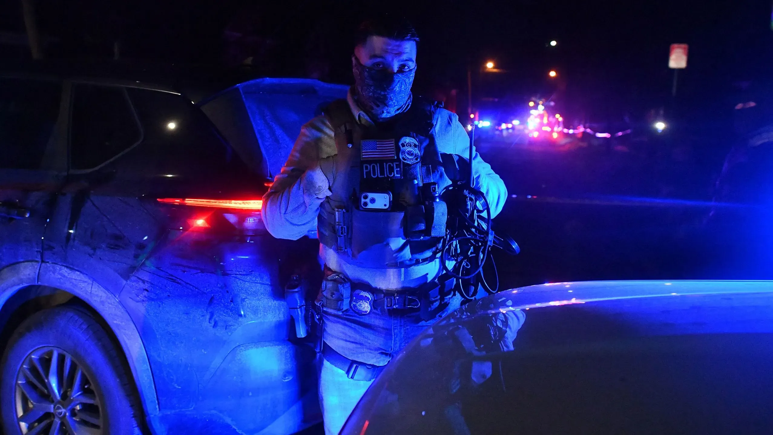 عميل من إدارة الهجرة والجمارك يحمل كاميرا هاتف محمول أثناء عملية في الليل، مع أضواء شرطة زرقاء في الخلفية.