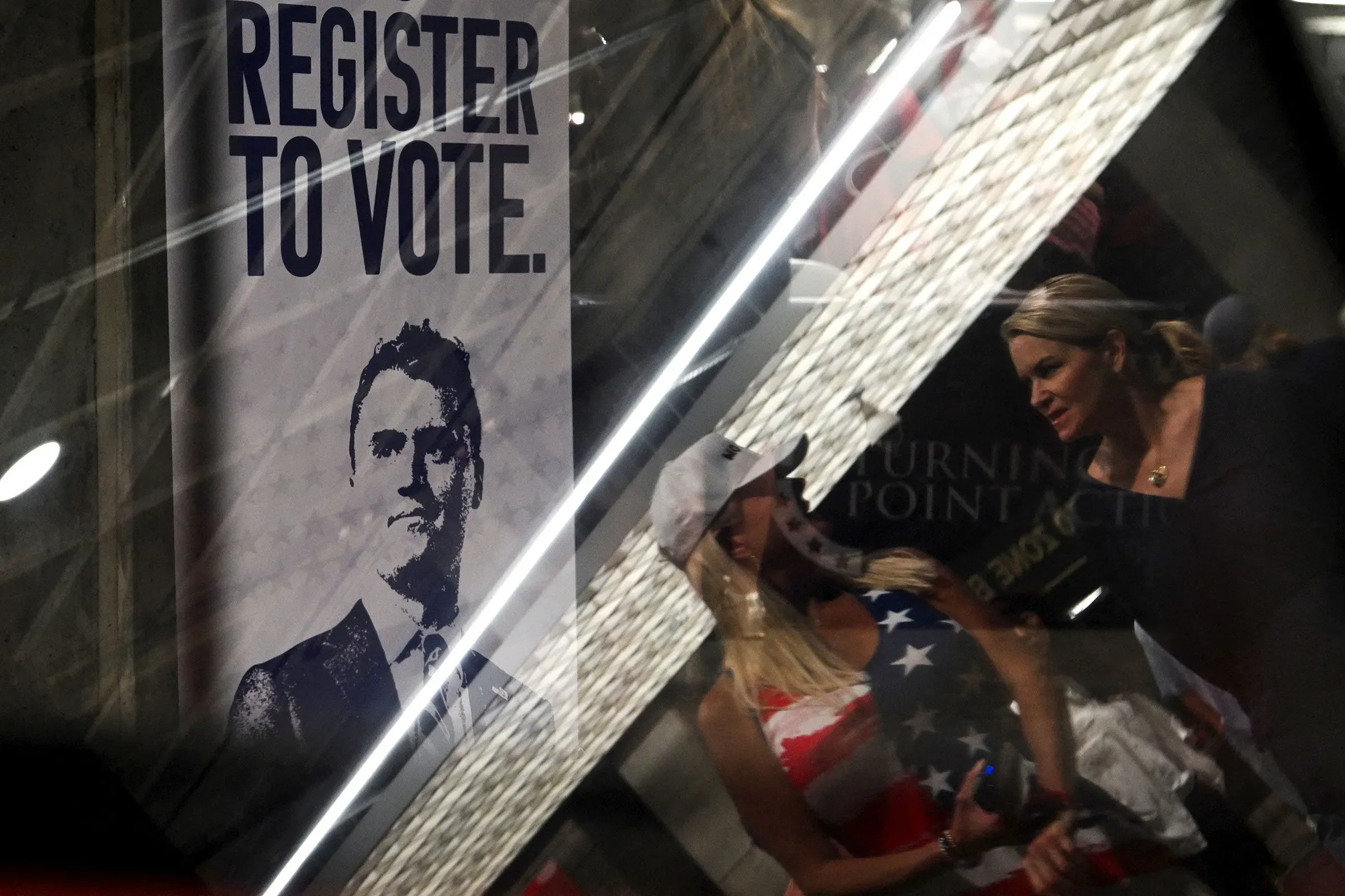 لافتة مكتوب عليها "سجل للتصويت" تظهر في حدث سياسي، مع أشخاص يتحدثون في الخلفية، تعكس أهمية المشاركة السياسية بعد مقتل تشارلي كيرك.