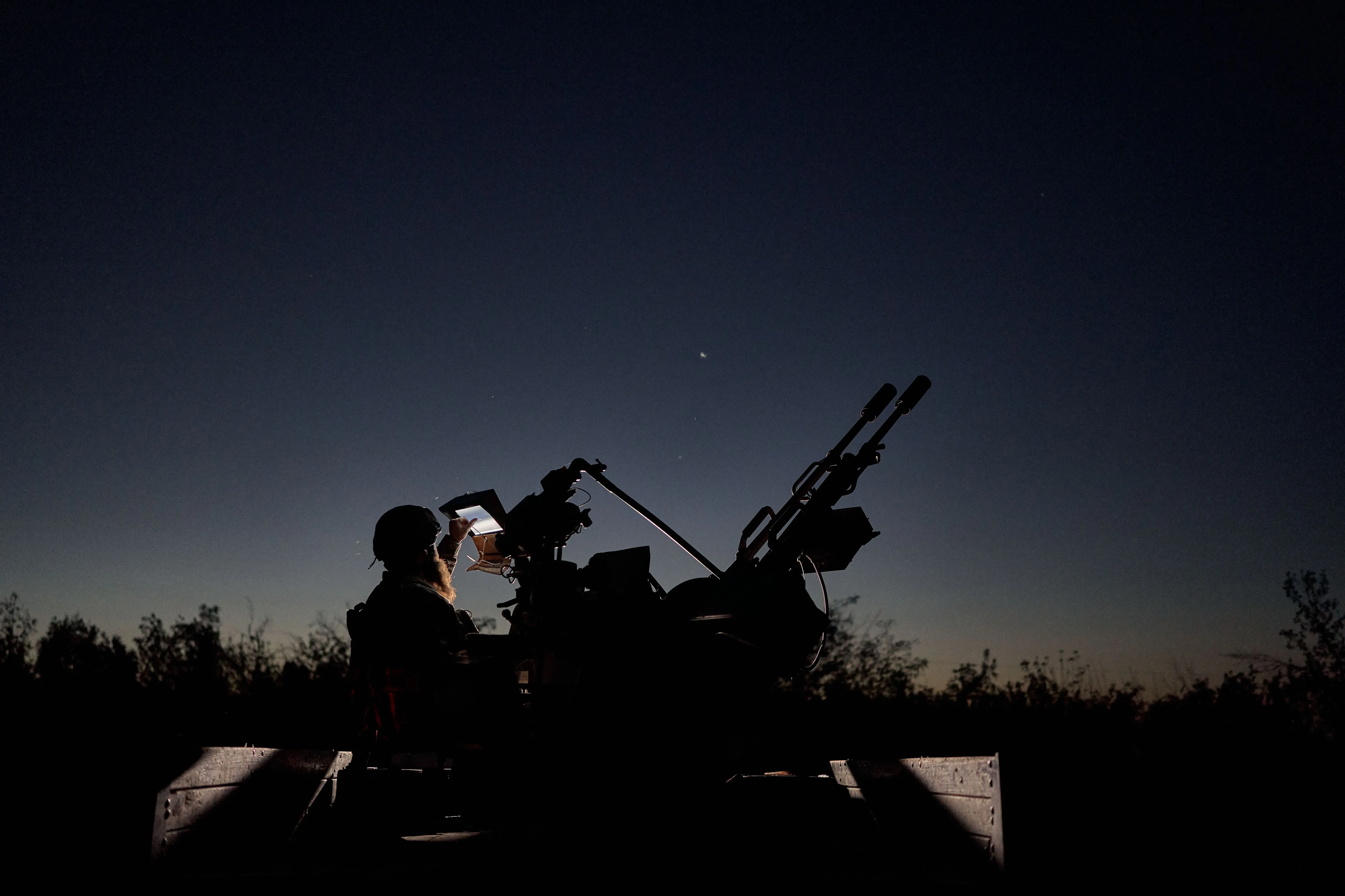 جندي أوكراني يجلس أمام مدفع مضاد للطائرات في الليل، يراقب السماء بحثًا عن أهداف محتملة، مع خلفية مظلمة تعكس التوتر في ساحة المعركة.