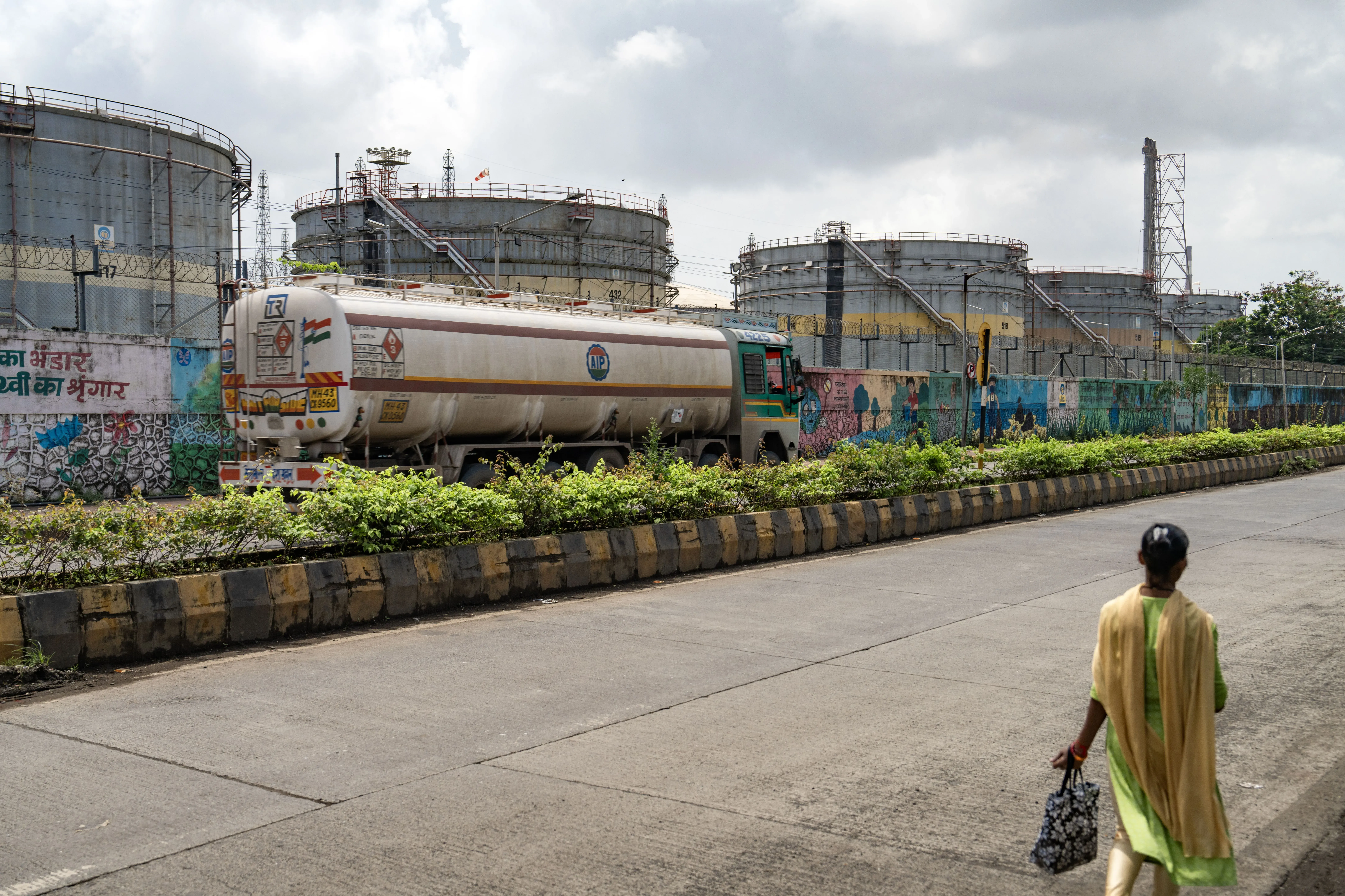 مشهد لامرأة تسير على الرصيف بجوار خزانات نفط كبيرة، مع شاحنة نقل تسير في الاتجاه المعاكس، مما يعكس النشاط الصناعي في المنطقة.