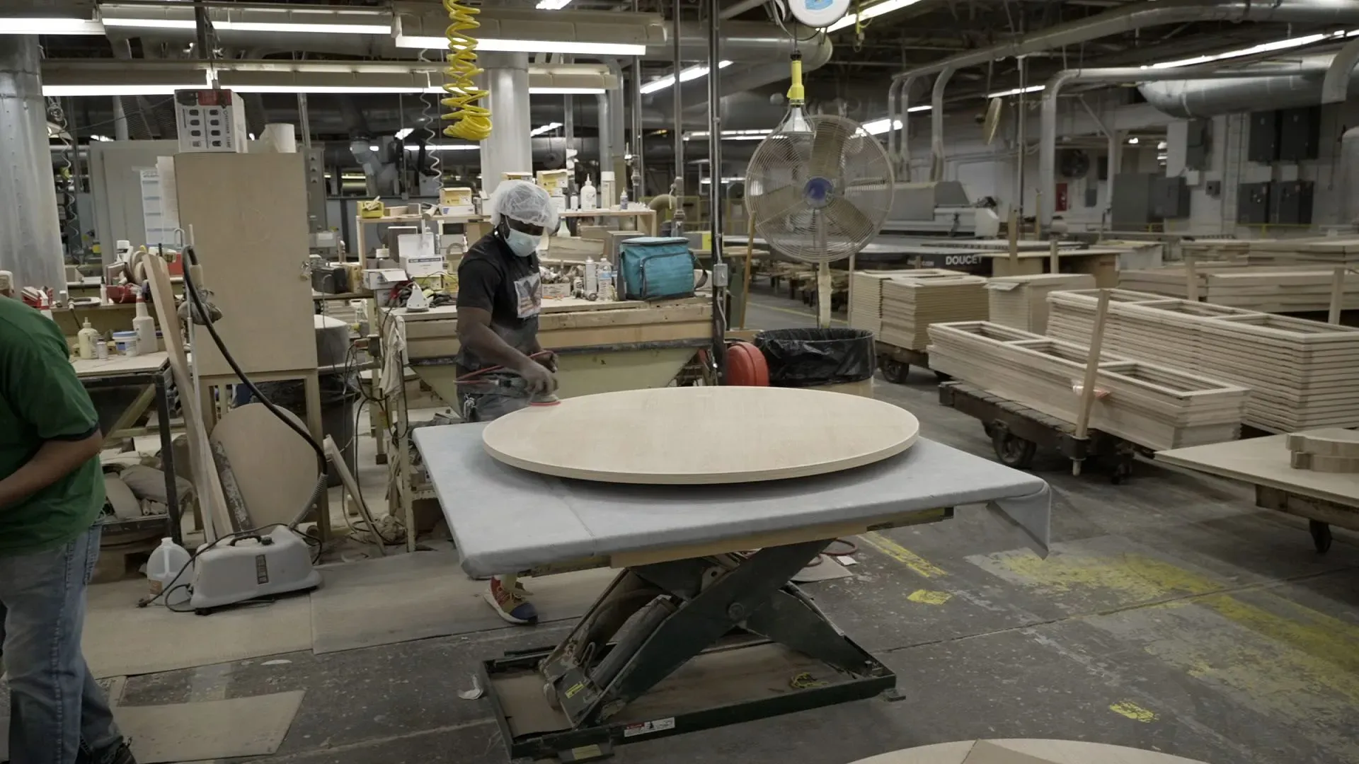 عامل في مصنع للأثاث يستخدم معدات لتجهيز قطع خشبية، مع وجود مواد خام وأثاث جاهز في الخلفية، مما يعكس جهود صناعة الأثاث في نورث كارولينا.