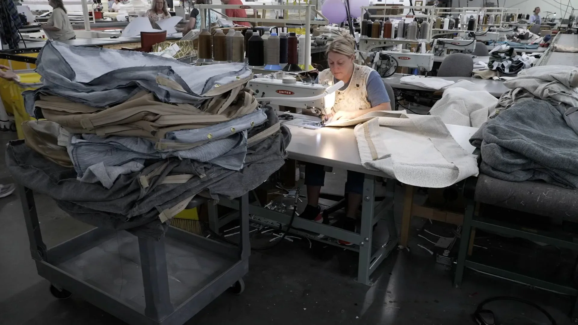 امرأة تعمل على ماكينة خياطة في مصنع أثاث، محاطة بأقمشة وأجزاء من الأثاث، تعكس جهود الصناعة في ولاية كارولينا الشمالية.