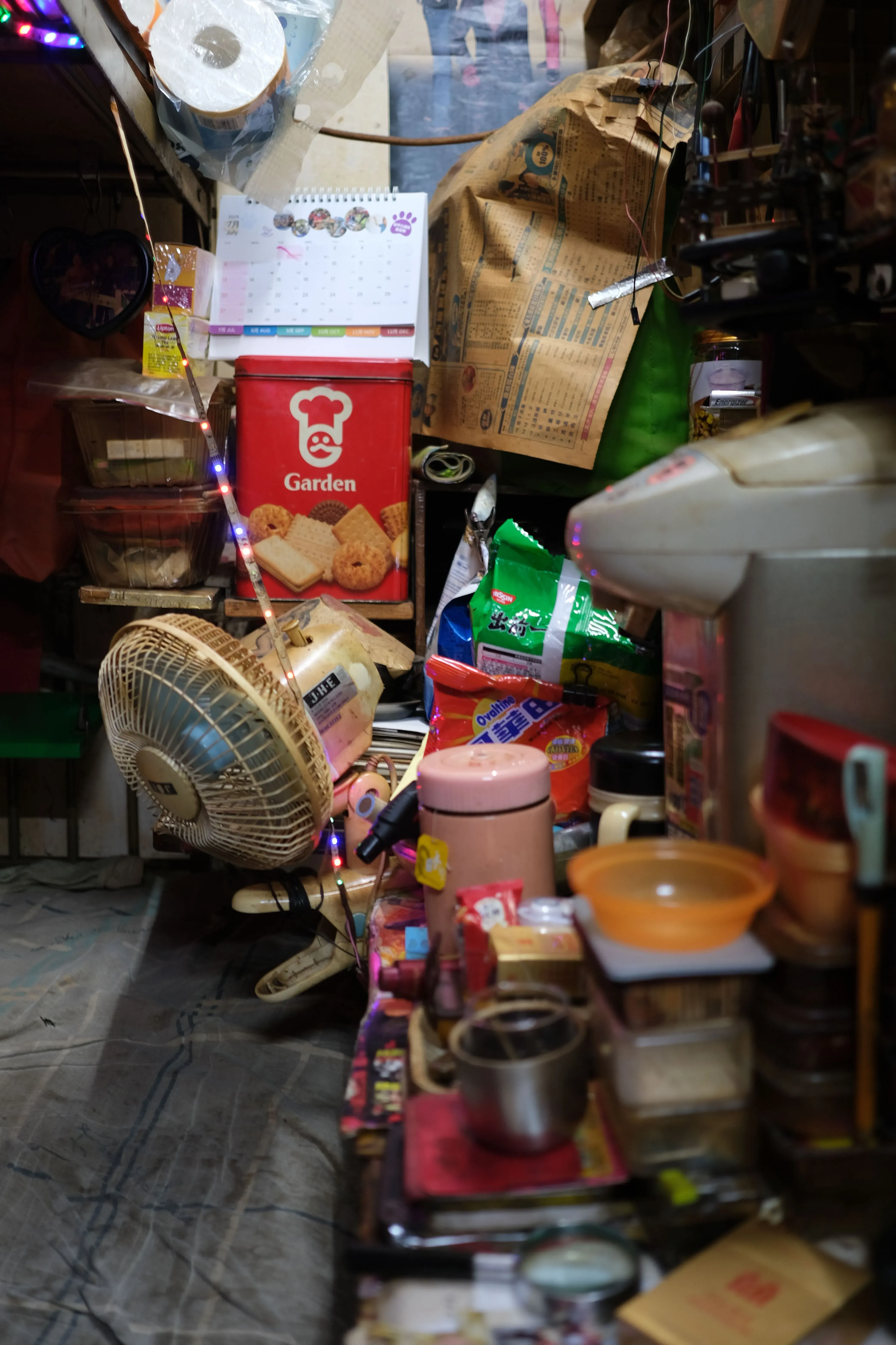 شقة صغيرة مزدحمة في هونغ كونغ، تحتوي على مروحة وأغراض منزلية، تعكس ظروف المعيشة الصعبة بسبب ارتفاع درجات الحرارة والرطوبة.