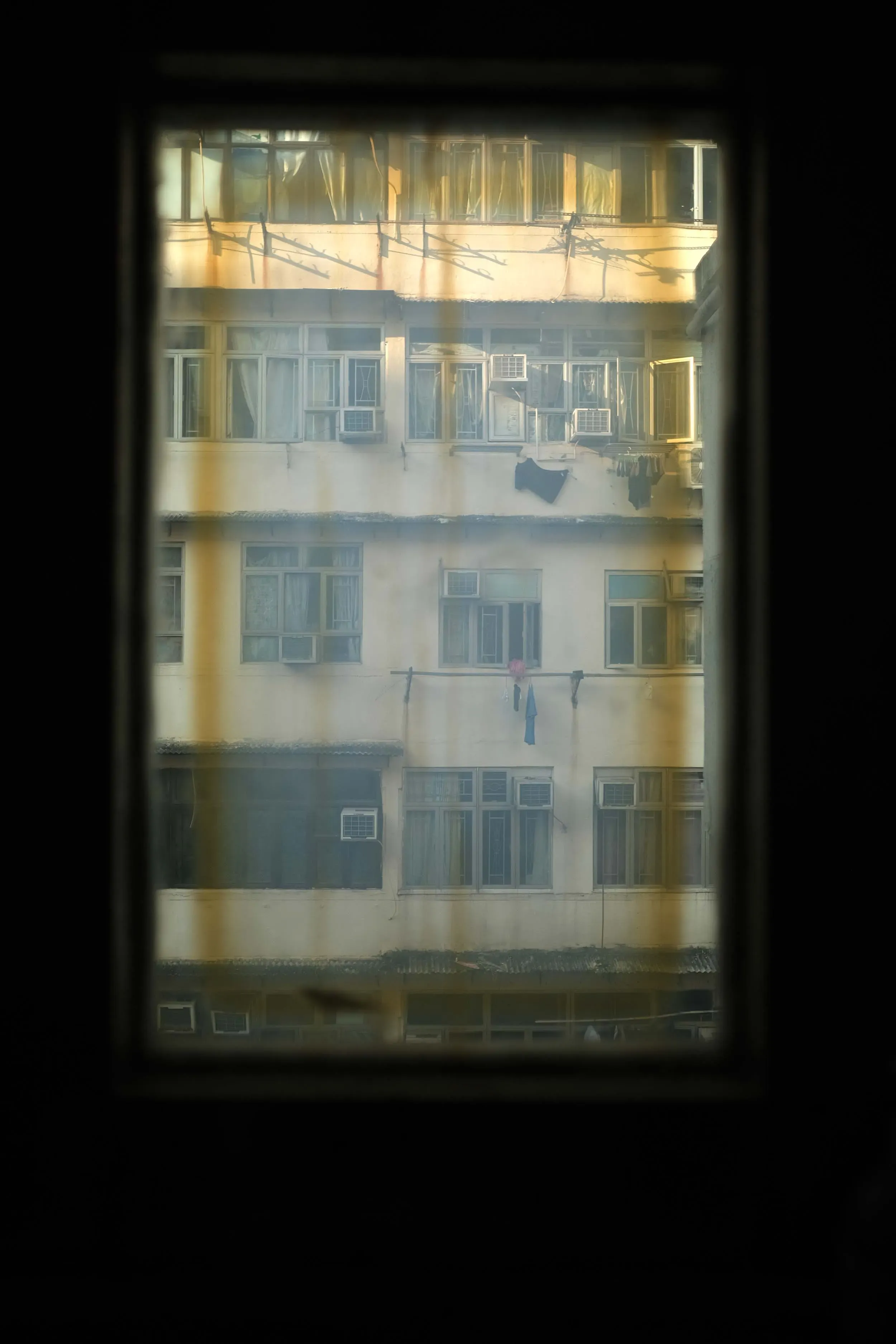 من خلال نافذة ضيقة، يظهر مبنى سكني في هونغ كونغ، حيث تتدلى الملابس وتظهر وحدات تكييف الهواء، مما يعكس ظروف المعيشة الصعبة في المدينة.