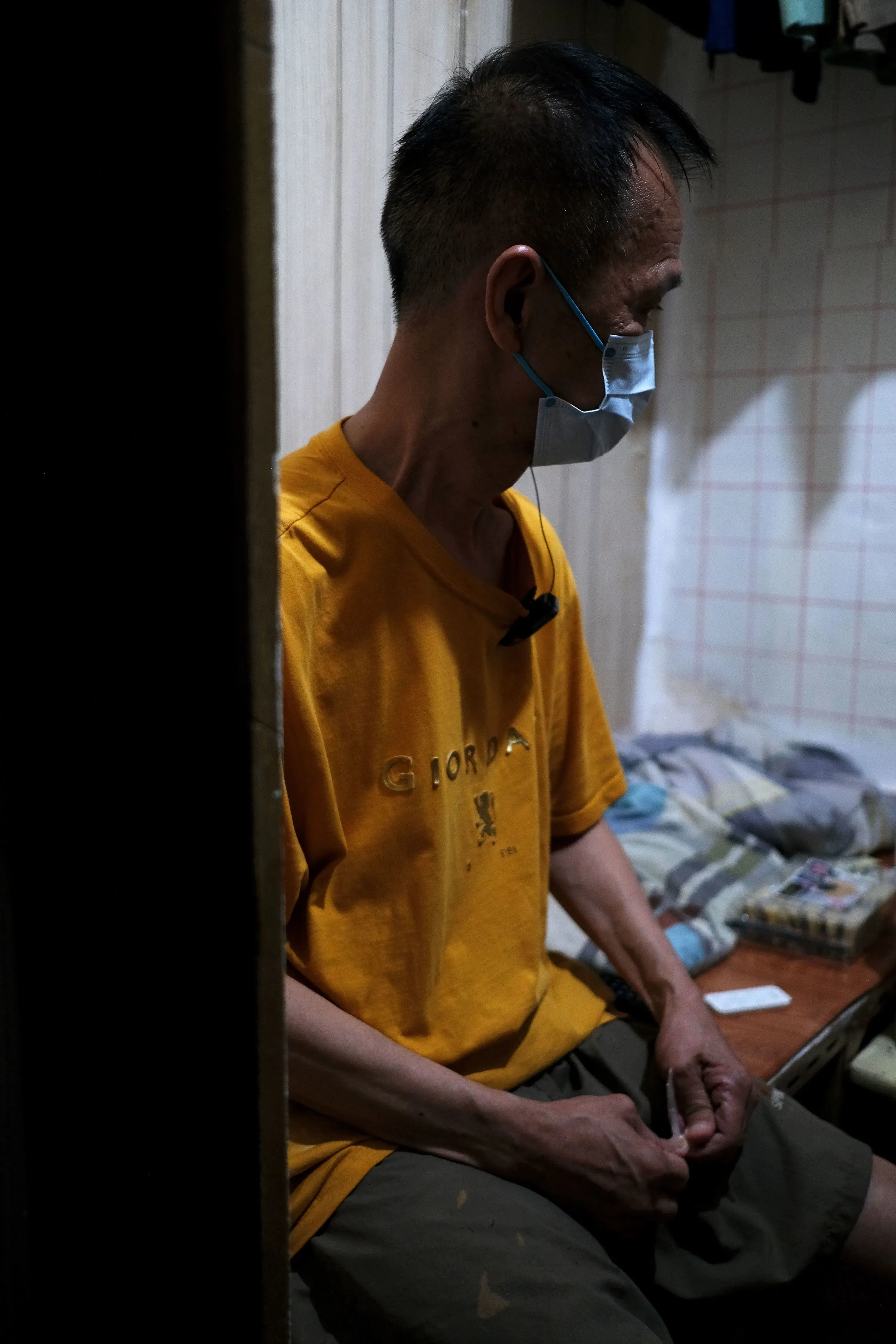 رجل مسن يرتدي قناع وجه، يجلس في غرفة ضيقة مع سرير صغير وأغراض مبعثرة، معبرًا عن ظروف المعيشة الصعبة في هونغ كونغ.