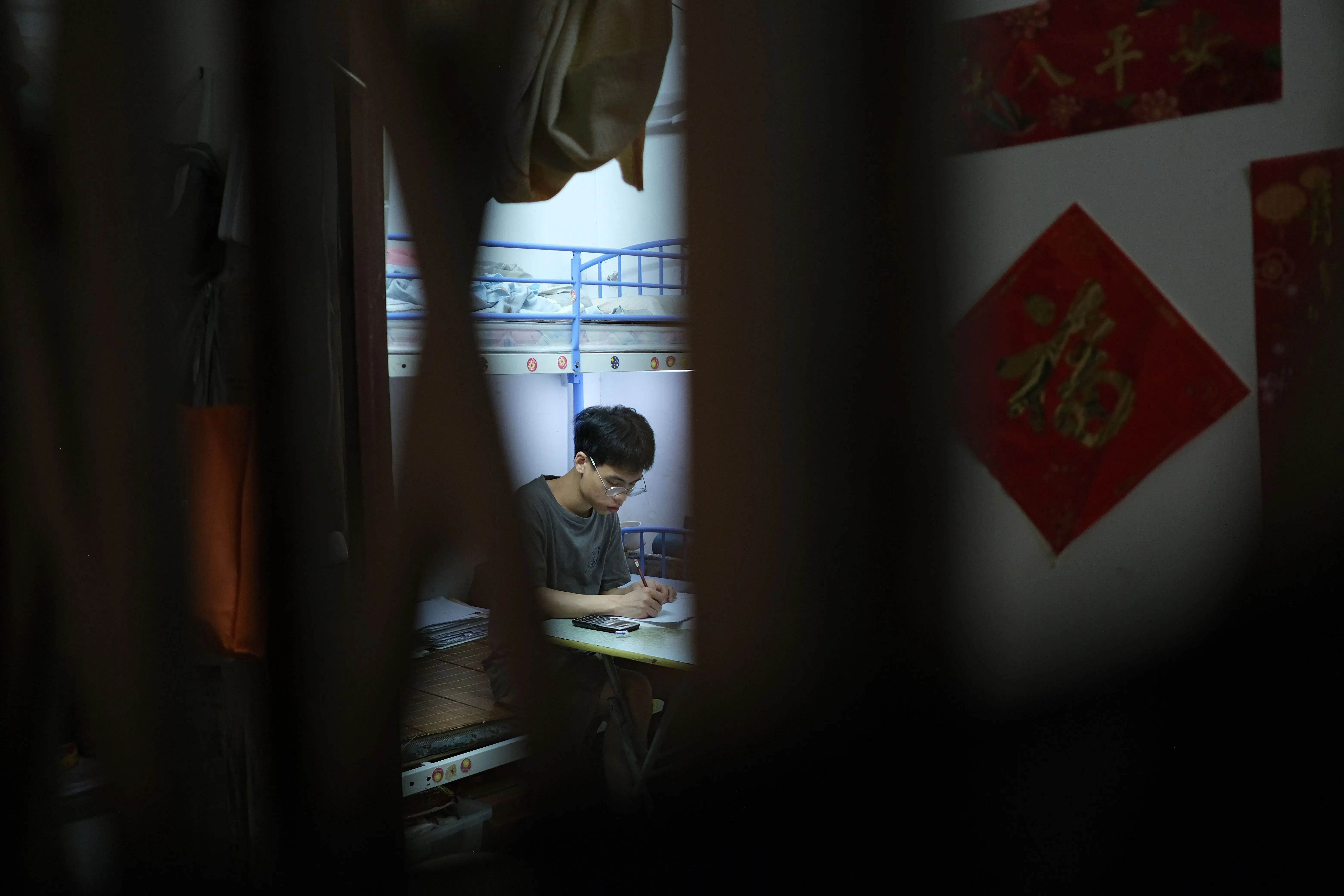 شاب يجلس على طاولة صغيرة في غرفة ضيقة، يكتب بتركيز. خلفه، أسرة متعددة الطوابق وزينة حائط تقليدية. تعكس الصورة ظروف المعيشة الصعبة في هونغ كونغ.