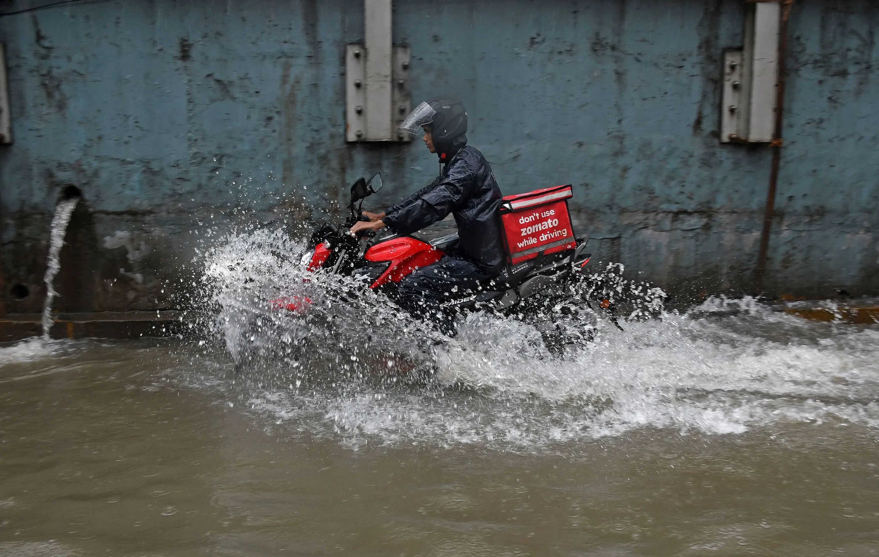 عامل توصيل يركب دراجة نارية في مياه راكدة، مرتديًا سترة واقية، مع شعار "لا تستخدم زوماتو أثناء القيادة" على حقيبته.