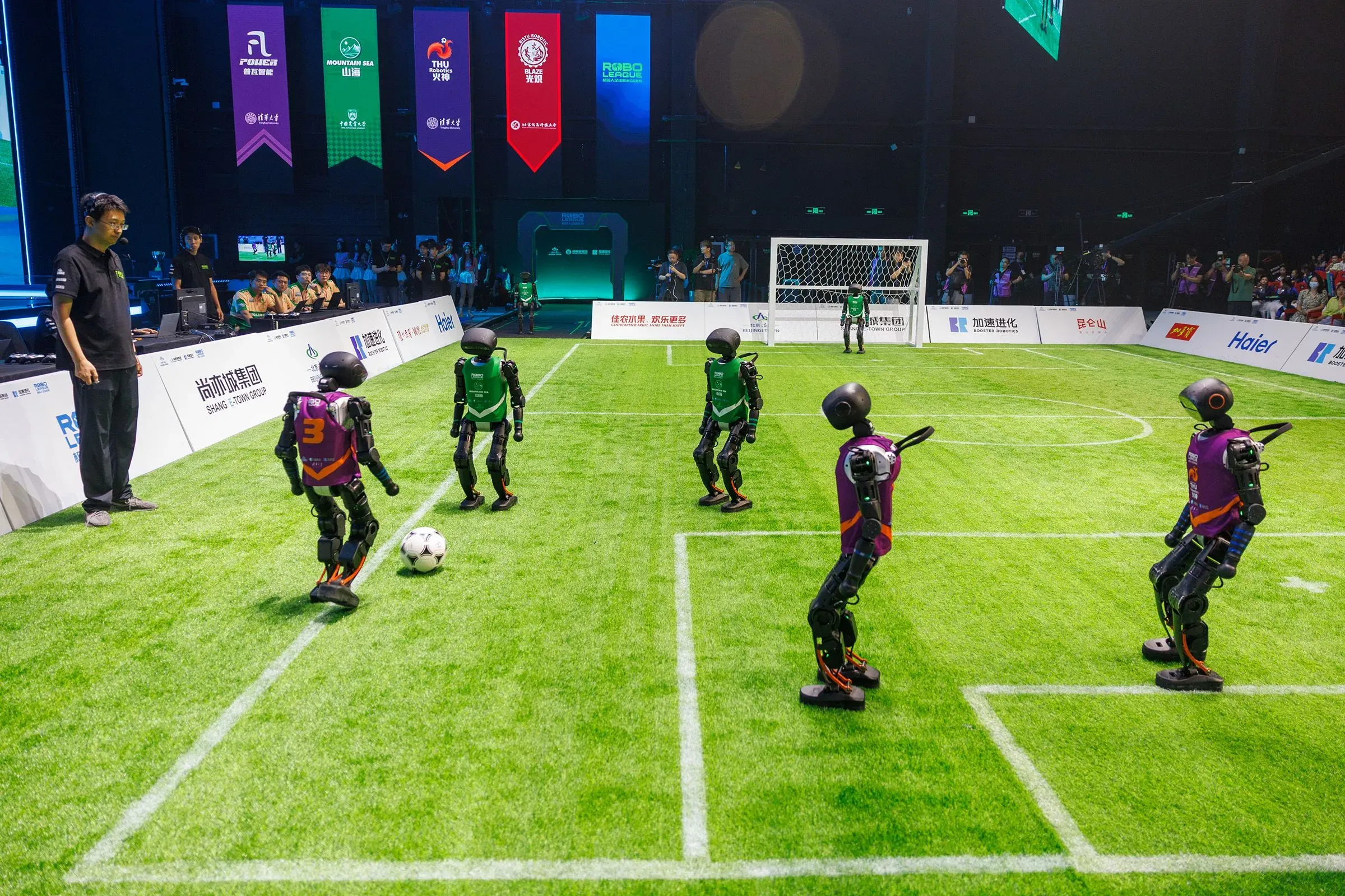 روبوتات تلعب كرة القدم في ملعب مخصص، مع وجود مدرب يراقب من الجانب. تعكس الصورة تقدم التكنولوجيا في الذكاء الاصطناعي.