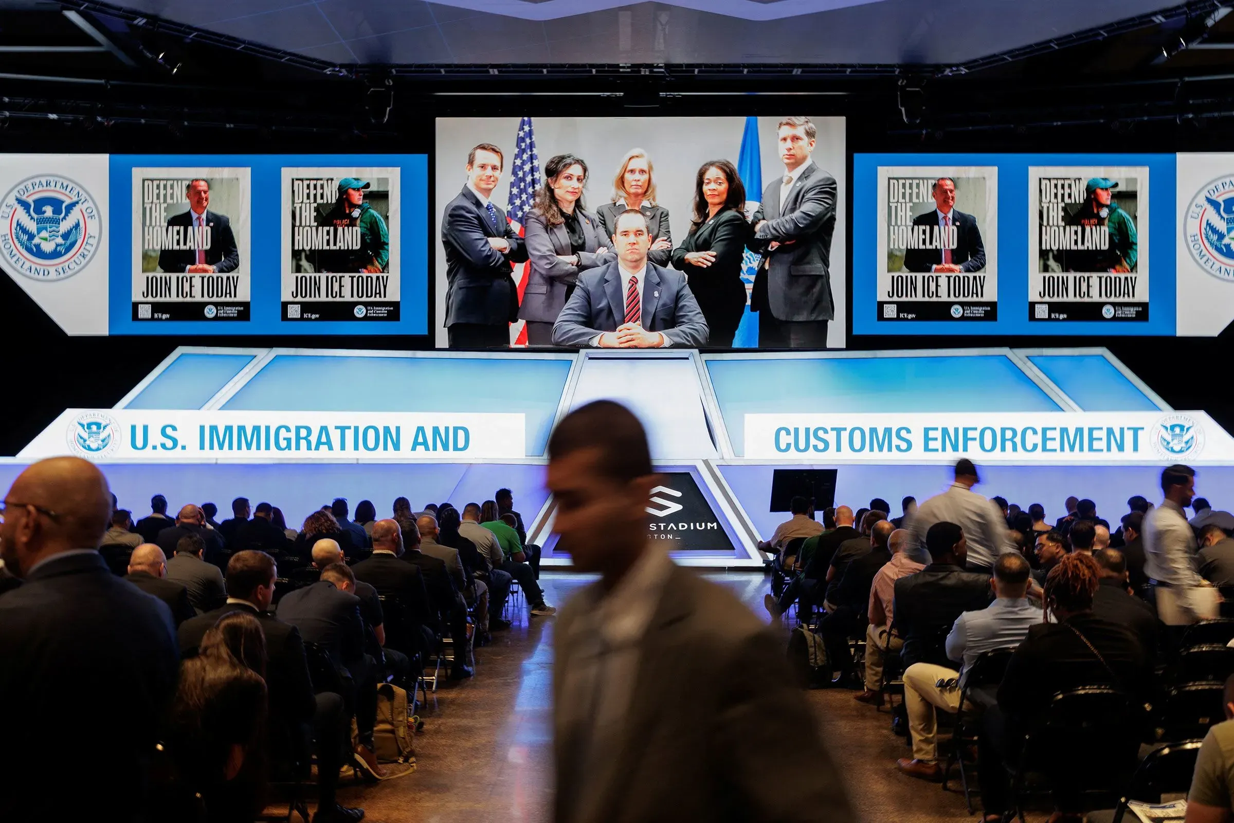 مؤتمر لوكالة إنفاذ قوانين الهجرة والجمارك، يظهر مسؤولون في مقدمة المسرح مع ملصقات تدعو للانضمام إلى الوكالة، وحضور كبير من المتقدمين.