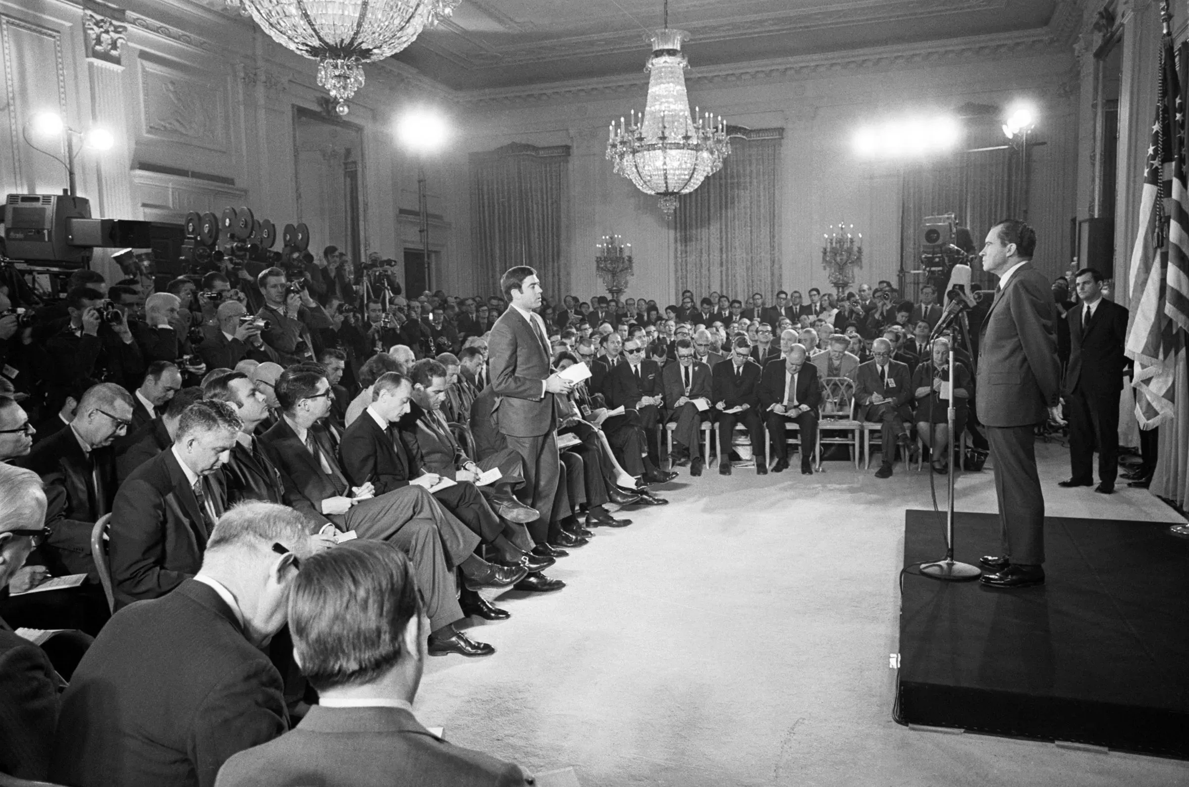 اجتماع صحفي في البيت الأبيض، حيث يتحدث الرئيس نيكسون أمام حشد كبير من الصحفيين، مع وجود كاميرات وإضاءة قوية.