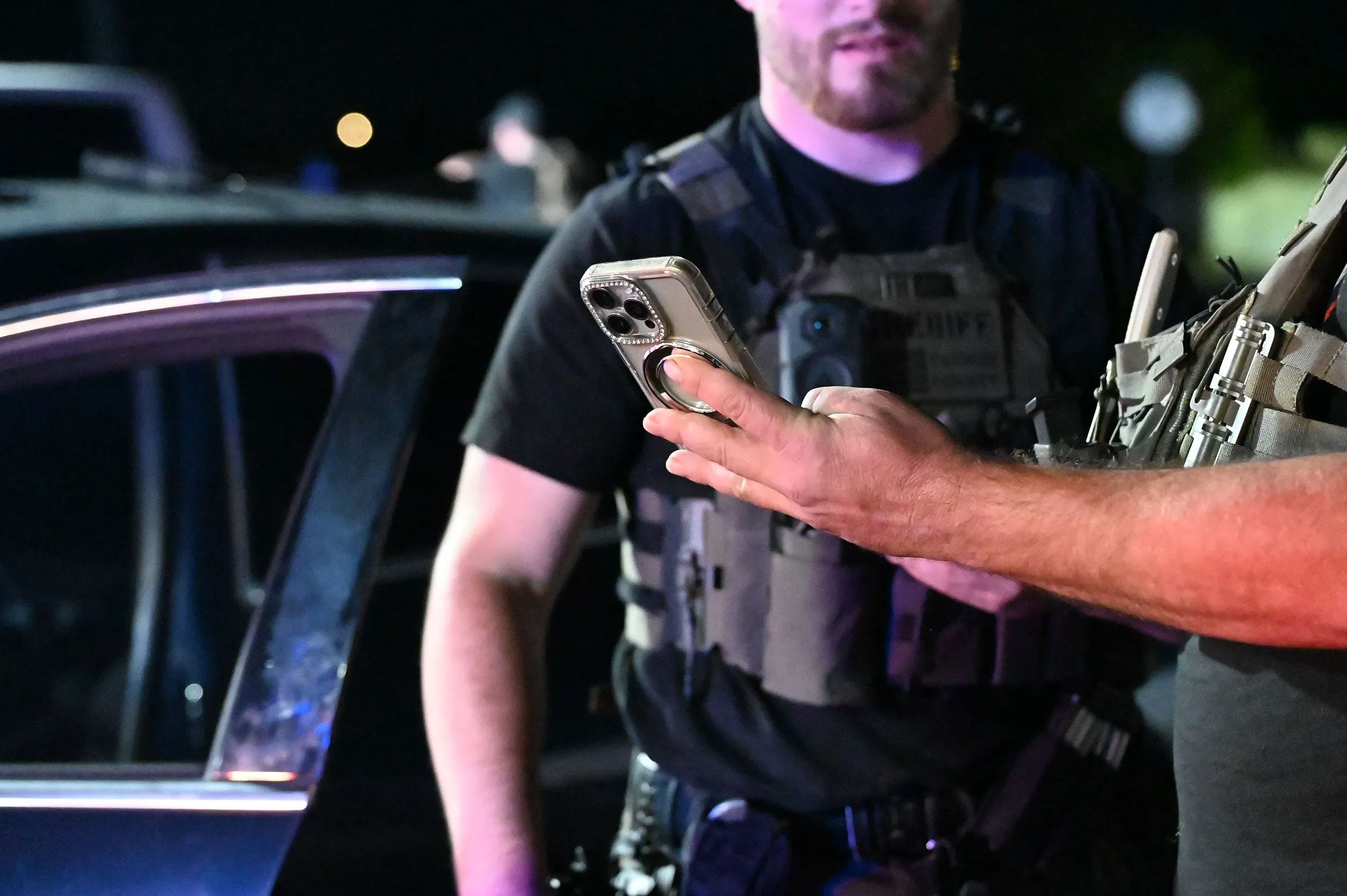 شرطي يحمل هاتفًا محمولًا أثناء عملية توقيف، مع وجود شرطي آخر في الخلفية، مما يعكس جهود مكافحة تهريب المهاجرين.