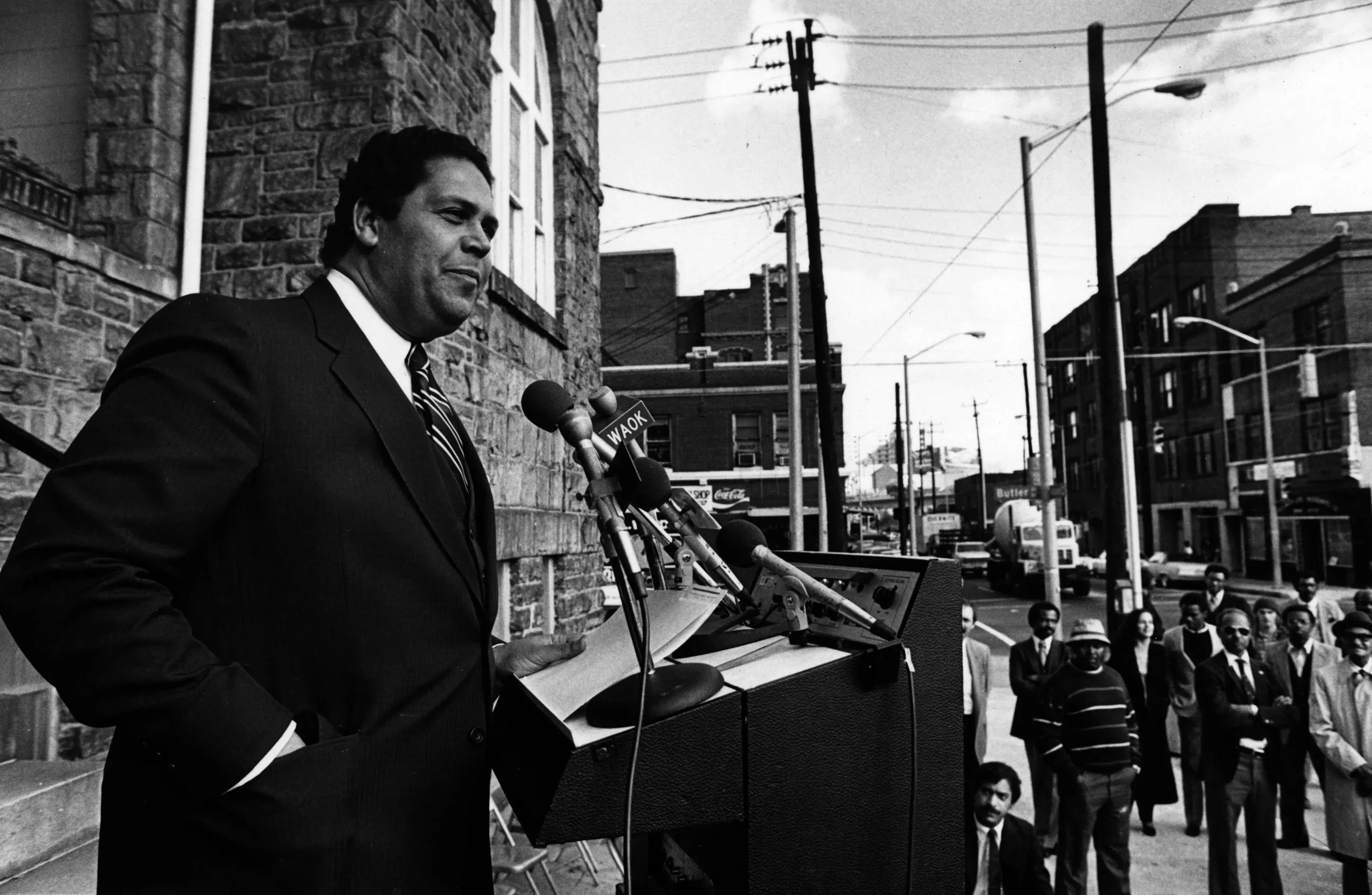 رجل يتحدث أمام ميكروفونات في مؤتمر صحفي، بينما يستمع له حشد من الناس في الشارع، مما يعكس قضايا التشرد والعدالة الاجتماعية.