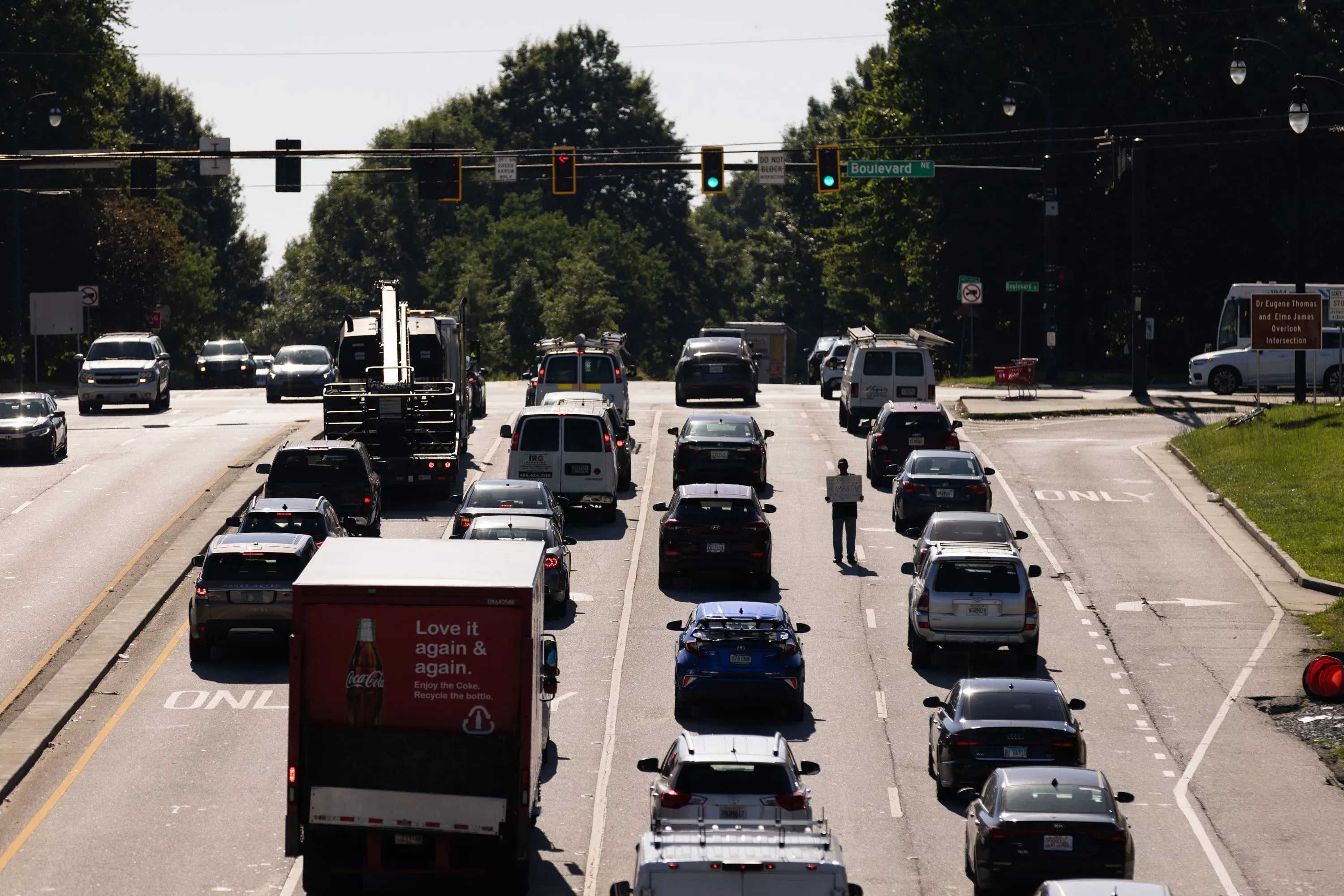 رجل يحمل لافتة "سأعمل مقابل الطعام" يقف في وسط شارع مزدحم، محاط بسيارات تسير ببطء، مما يعكس تحديات التشرد في المجتمع.