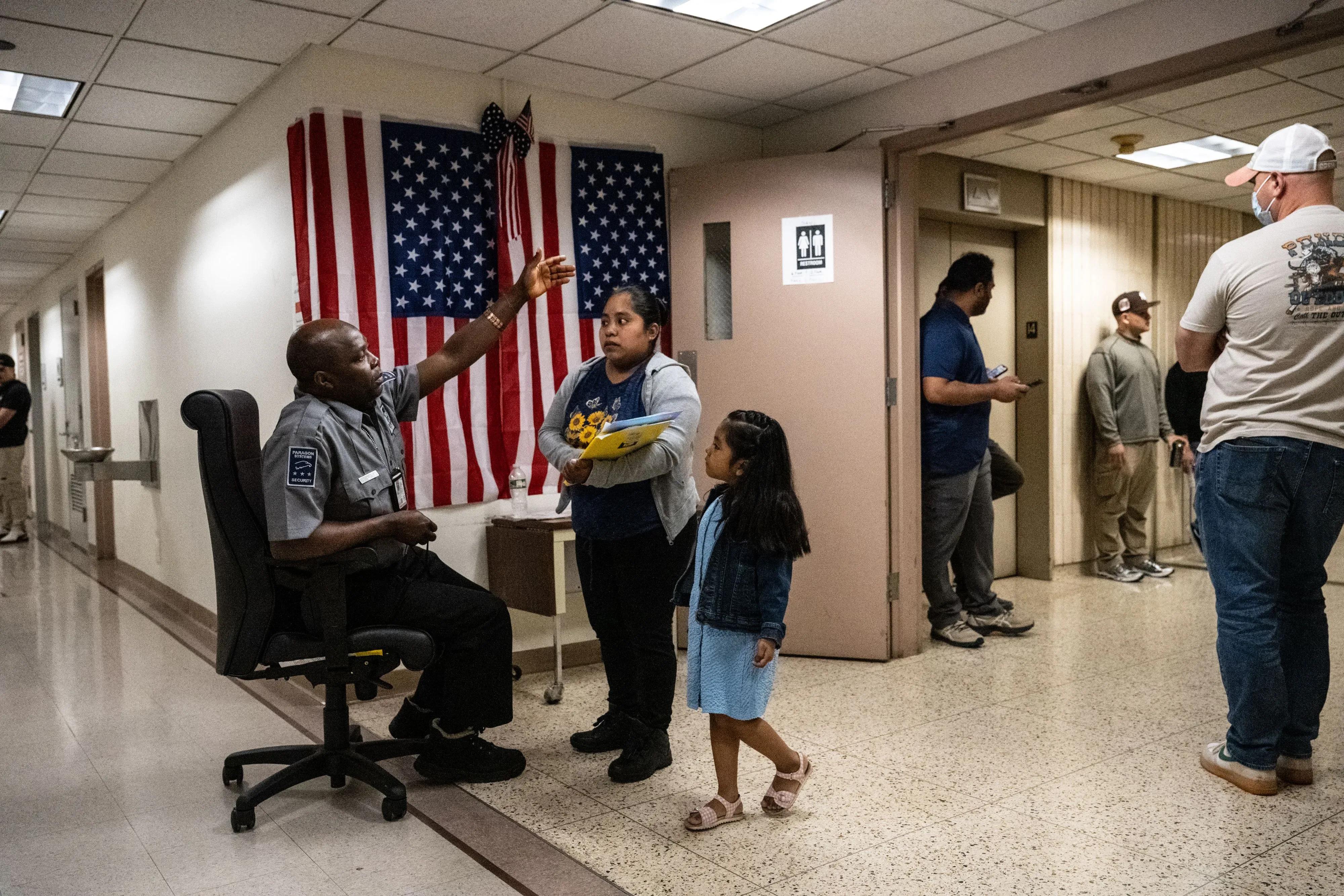 امرأة تحمل أوراقًا وتقف مع طفلة صغيرة أمام موظف أمني في قاعة محكمة، مع وجود العلم الأمريكي في الخلفية، مما يعكس أجواء قضايا الهجرة.