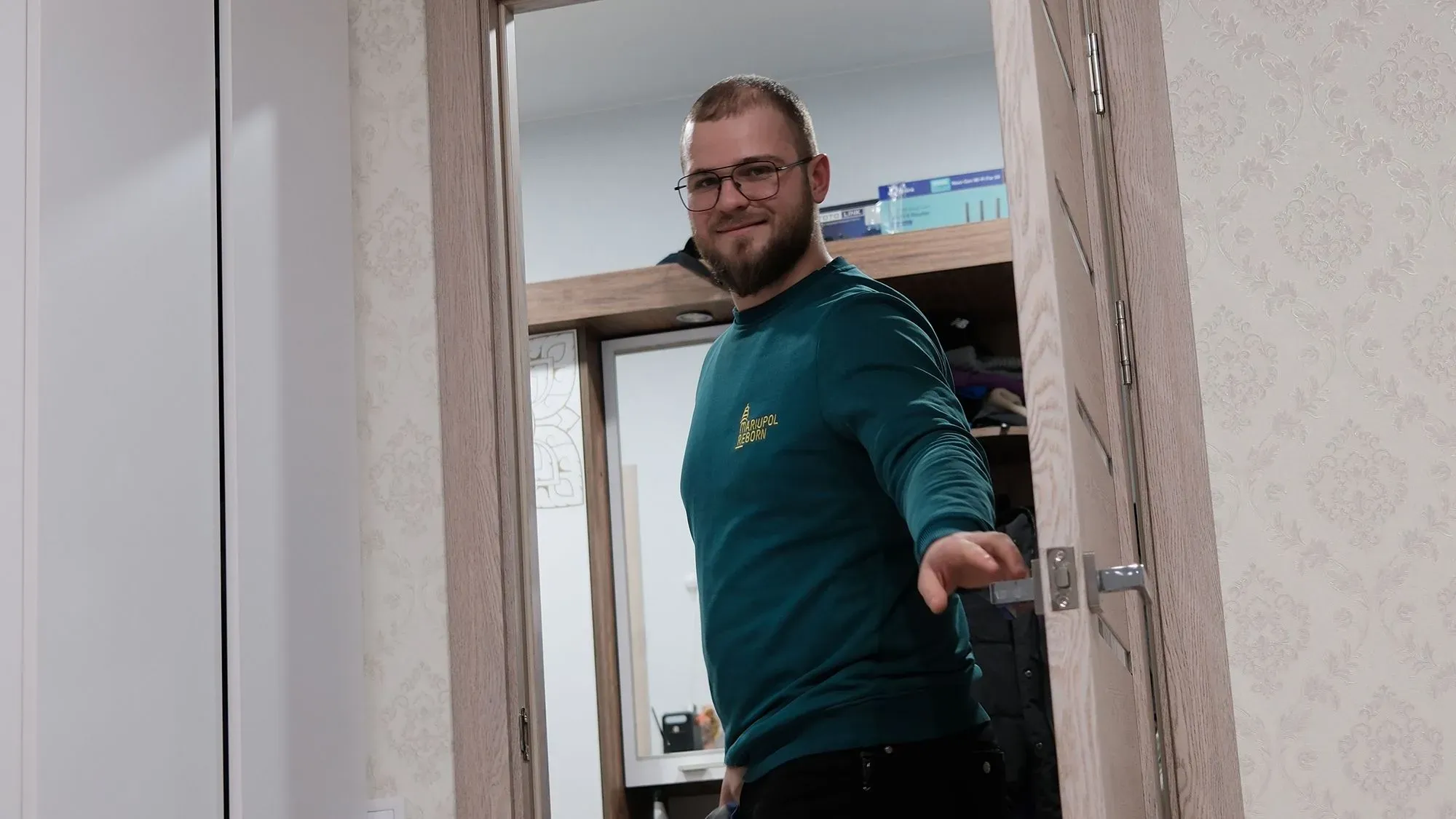 شاب مبتسم يقف عند باب غرفة، يرتدي سترة زرقاء، ويظهر في الخلفية أثاث منزلي، مما يعكس حياة جديدة في أوكرانيا بعد العودة.