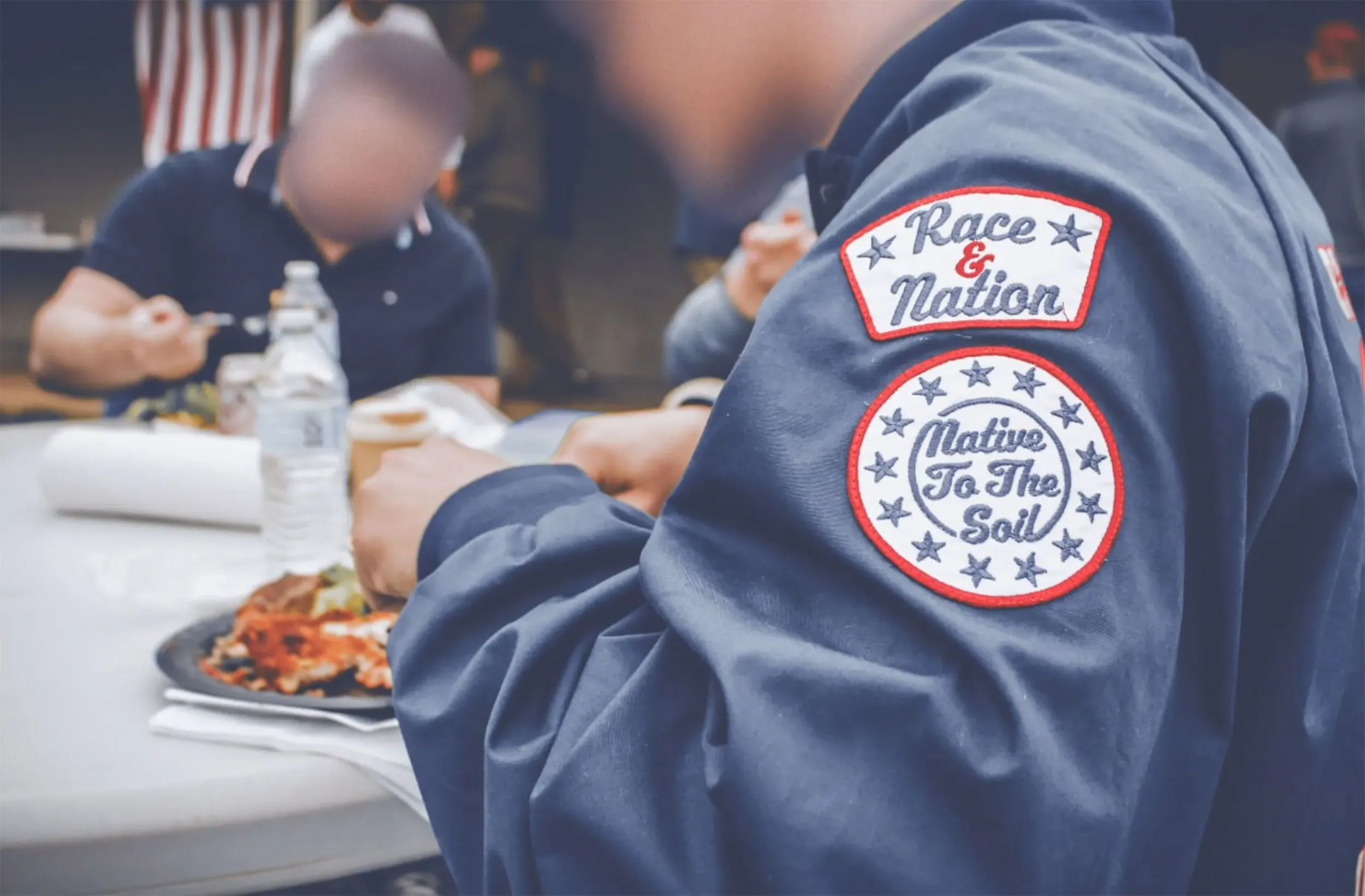 شخص يرتدي سترة تحمل شعارات تتعلق بالعرق والأمة، بينما يتناول الطعام مع مجموعة من الأشخاص في مناسبة اجتماعية.