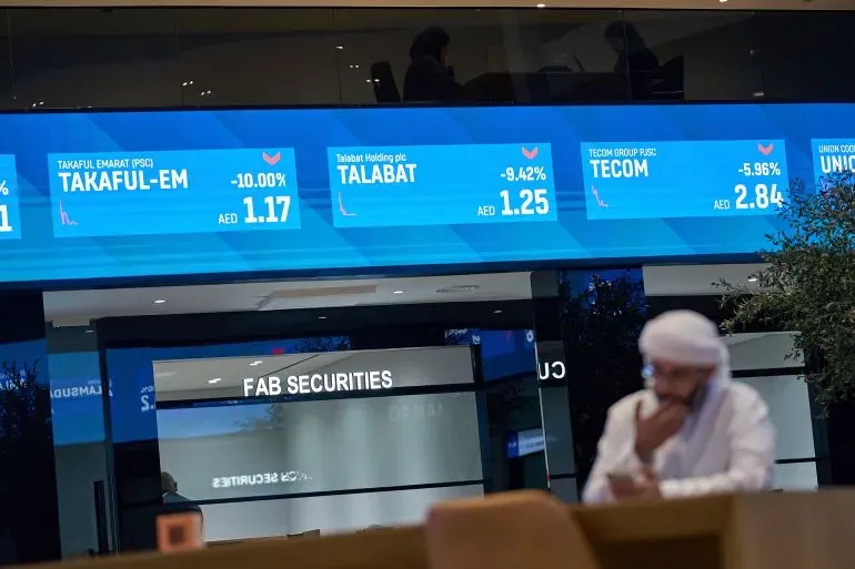 تظهر الشاشة في سوق الأسهم الإماراتي انخفاضًا في أسعار الأسهم، مع بيانات عن شركات مثل "تأمين الإمارات" و"طلبات"، بينما يجلس مستثمر يتابع السوق.
