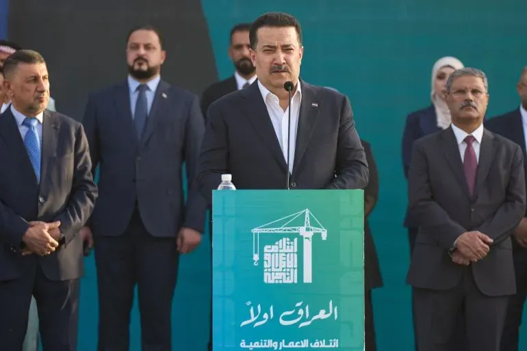رئيس الوزراء العراقي محمد شياع السوداني يتحدث خلال فعالية انتخابية، محاطًا بعدد من المسؤولين، مع شعار تحالف البناء والتنمية خلفه.
