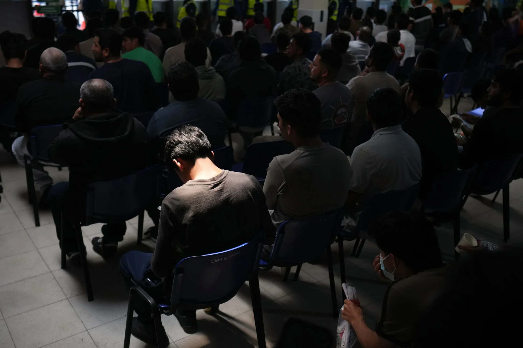 مجموعة من المهاجرين ينتظرون في مركز احتجاز مظلم، يعكس حالة القلق وعدم اليقين في ظل سياسة ترحيل المهاجرين إلى دول غير معروفة.