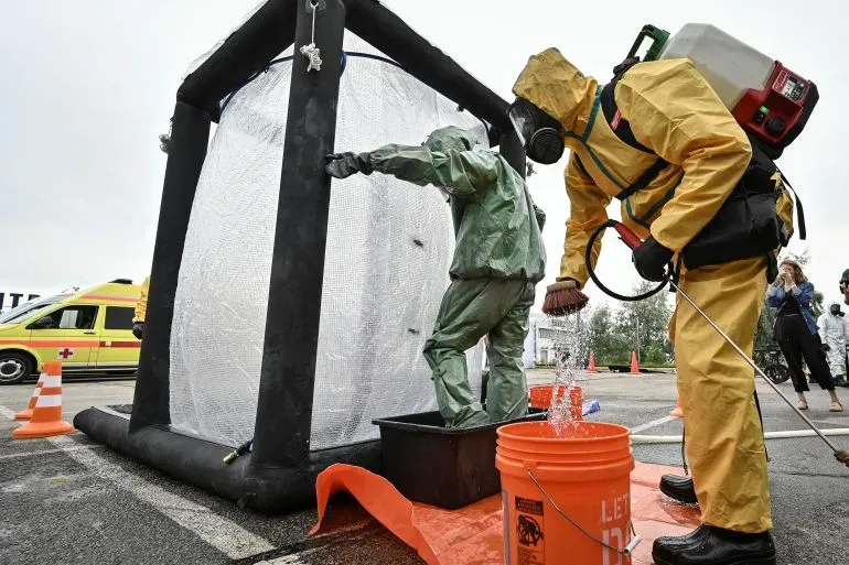 أفراد يرتدون ملابس واقية يقومون بإجراء عملية تعقيم في موقع طوارئ، مع وجود خزان مياه وصناديق في الخلفية، مما يعكس الاستعدادات لمواجهة الحوادث النووية.