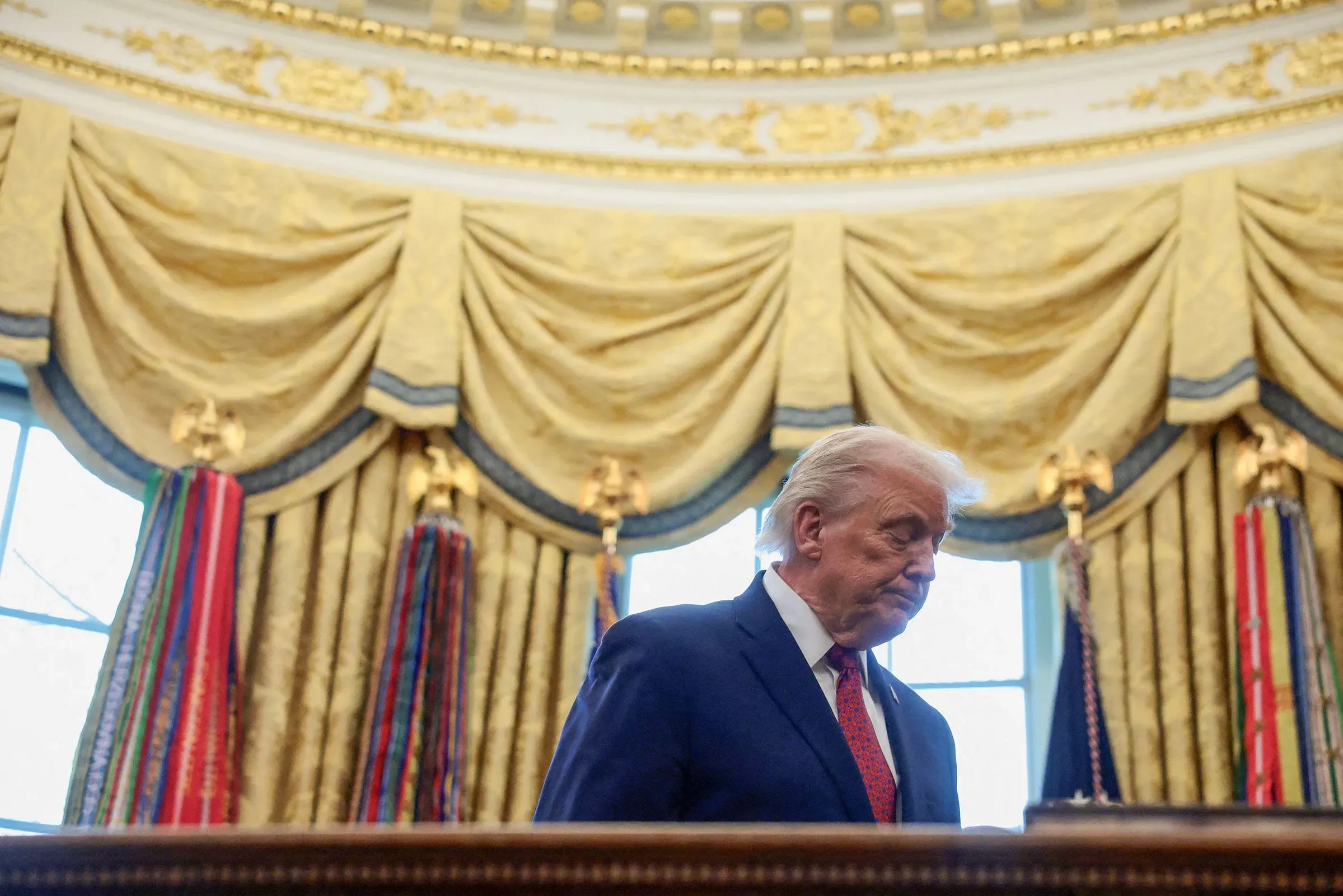 ترامب يسير في المكتب البيضاوي، يبدو متجهمًا، محاطًا بستائر ذهبية وميداليات، في لحظة تعكس الضغوط السياسية المتزايدة.