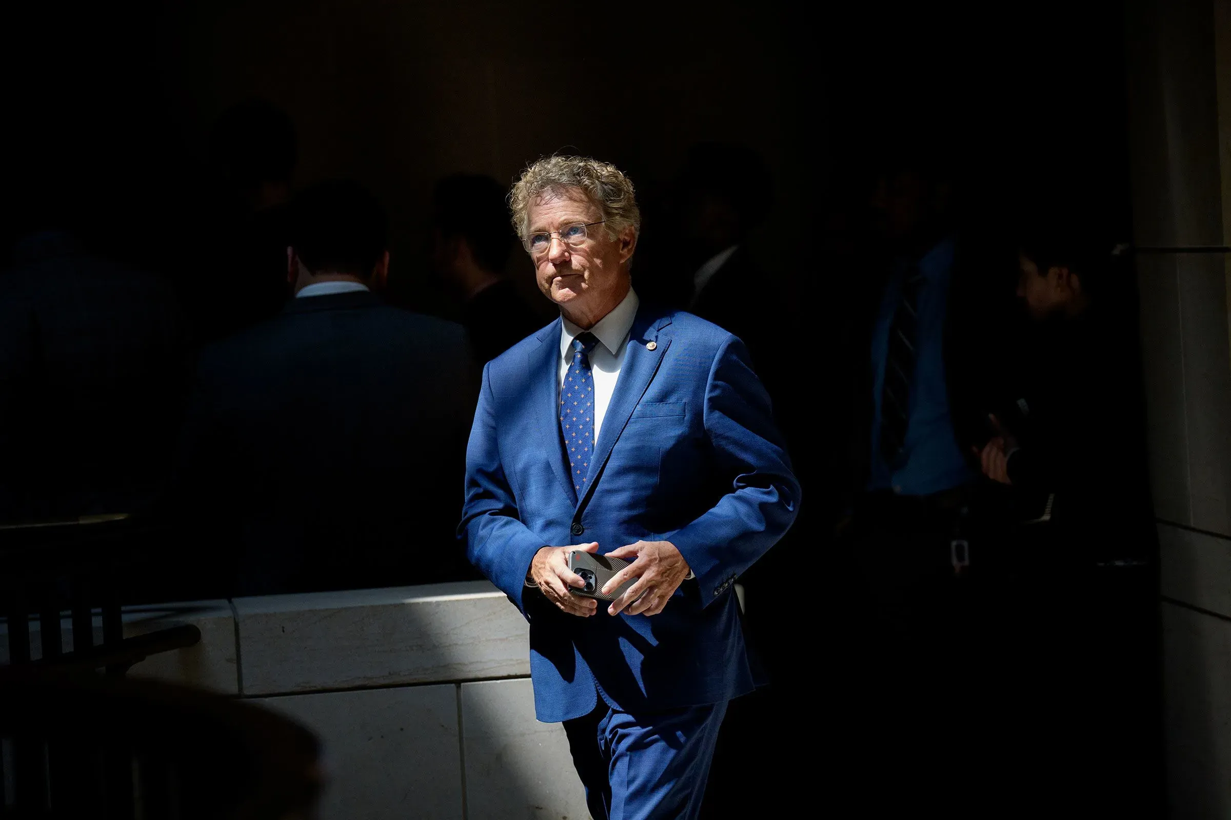 سيناتور إنديانا جين ليسينج يرتدي بدلة زرقاء، يسير في ممر مضاء بشكل خافت، مع وجود أشخاص آخرين في الخلفية، معبرًا عن معارضته لسياسات ترامب.