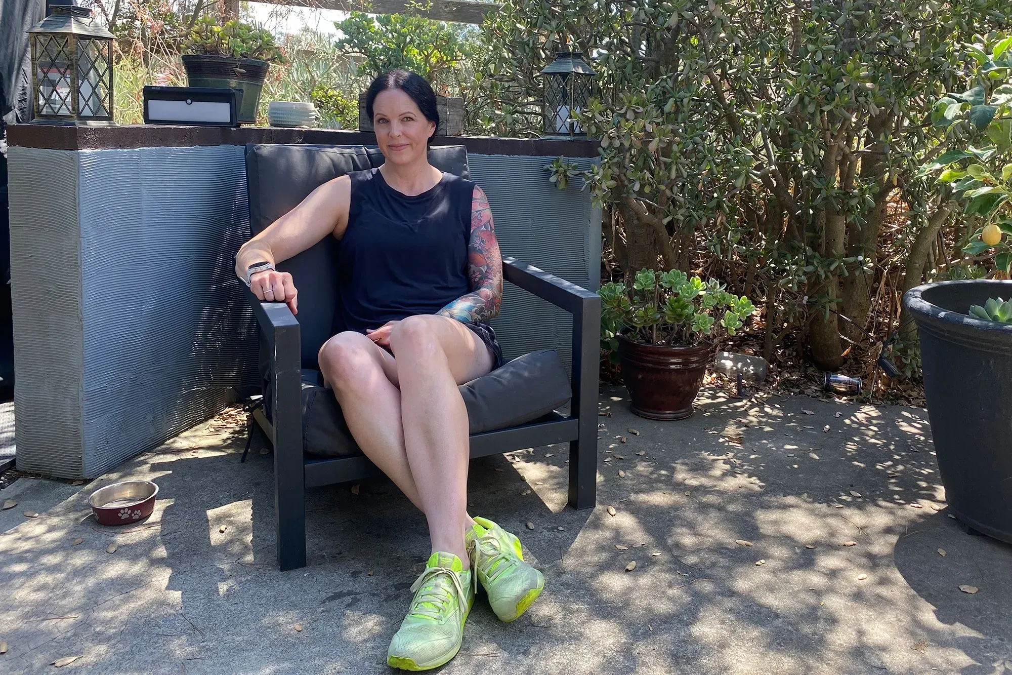 كاتي كوريو تجلس في حديقة، مرتدية قميصاً أسود وسروالاً قصيراً، مع أحذية رياضية زاهية، تعبر عن تجربتها في إزالة غرسات الثدي.