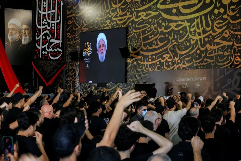 تجمع حشود من أنصار حزب الله في قاعة مزينة بشعارات الحزب، حيث يُعرض خطاب الأمين العام حسن نصر الله على شاشة كبيرة.