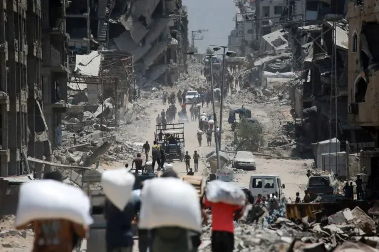 تظهر الصورة مشهدًا من غزة حيث يسير فلسطينيون حاملين أكياسًا من المساعدات في منطقة مدمرة، وسط الدمار الناتج عن الصراع المستمر.