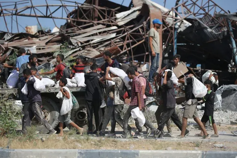 مجموعة من الأطفال والشبان يحملون أكياسًا من الطعام بالقرب من مباني مدمرة في غزة، وسط أزمة إنسانية متفاقمة.