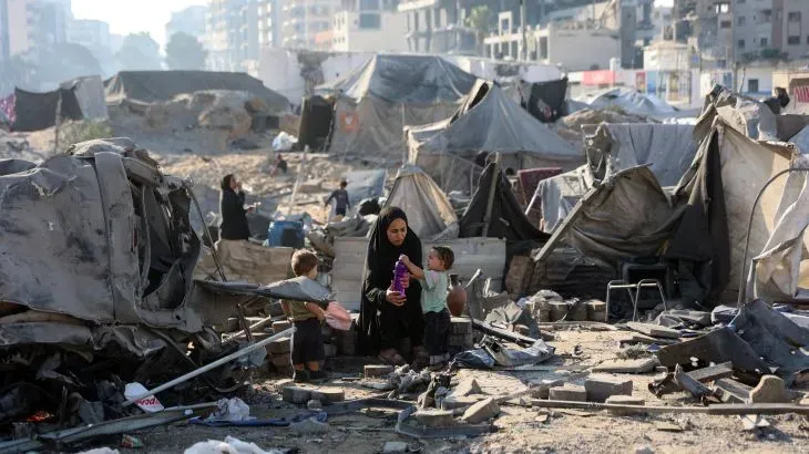 امرأة ترتدي الحجاب تجلس مع طفلين وسط خيام مهجورة وأنقاض في مدينة غزة، تعكس معاناة النازحين في ظل الأوضاع الصعبة.