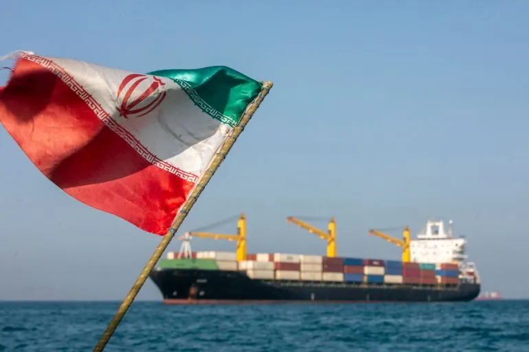 علم إيران يرفرف في المقدمة، بينما يظهر في الخلفية سفينة شحن كبيرة، مما يرمز إلى النفوذ الإيراني في التجارة البحرية.