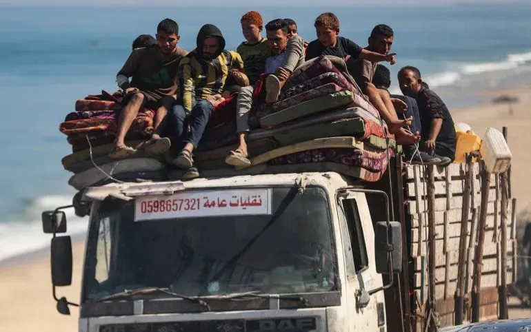 مجموعة من الشباب يجلسون على ظهر شاحنة محملة بالمفروشات، بينما يتجهون نحو منطقة آمنة على شاطئ غزة، وسط أجواء من النزوح.