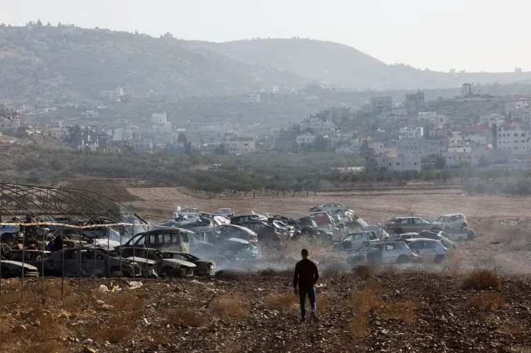 صورة لشاب يسير في أرض زراعية بالقرب من مخيم للاجئين، محاطة بسيارات مدمرة، تعكس آثار التهجير في الضفة الغربية.