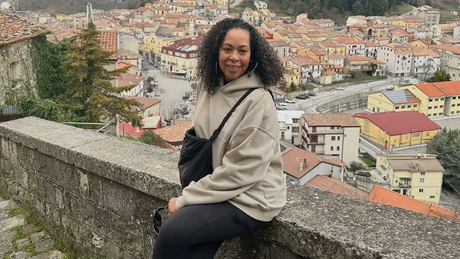 امرأة مبتسمة تجلس على حافة جدار حجري، خلفها منظر جميل لقرية لاترونيكو الإيطالية مع منازل ملونة وجبال.
